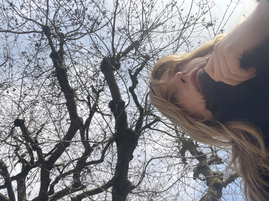 A low-angle, upward-facing photographic image depicts the upper portion of a light-skinned individual with long blonde hair, seen from below, against a backdrop of a large, bare tree and a bright sky. The individual, an adult female, is positioned in the lower-left quadrant, looking upwards and to the right. Her right hand is visible, gripping the dark-colored collar of her garment, partially covering her chin and lower mouth. A small silver stud is present in her right nostril. Her lips are a soft pink. The upper two-thirds of the frame are dominated by the dark, intricate network of gnarled, leafless branches of a large deciduous tree, consistent with a winter season. Small, dark, spherical objects, likely berries or seed pods, cling to some finer branches. The sky behind the tree is bright, a pale blue with diffuse white areas. Sunlight appears to illuminate the scene from the upper-right, casting highlights on the individual's hair and backlighting the tree branches. The environment is consistent with an outdoor public space, such as a park or tree-lined street, commonly found in London, United Kingdom.