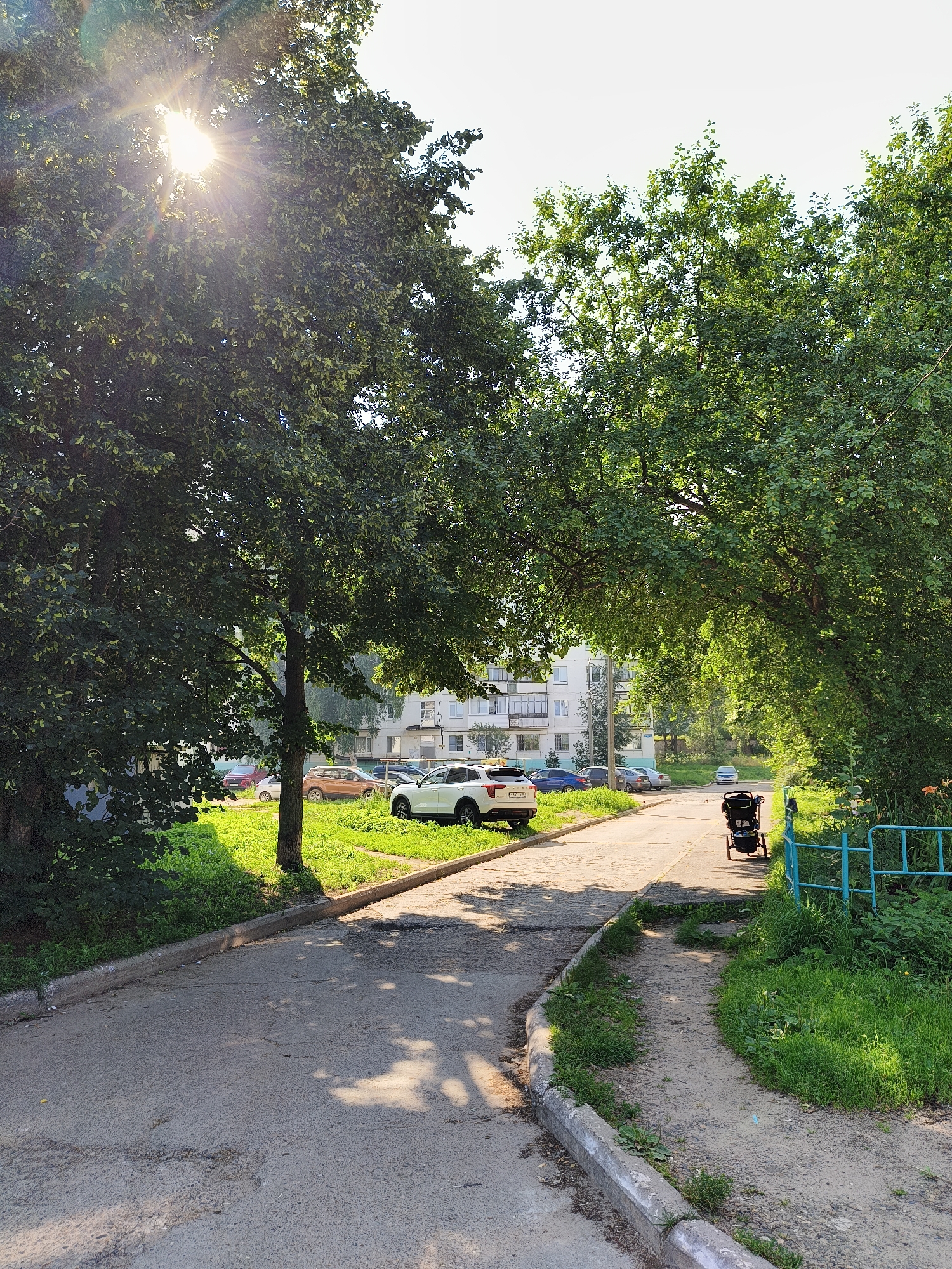 The image depicts a sun-drenched, tree-lined residential street scene. Sunlight streams through the lush green canopy of mature trees, casting dappled shadows on the paved road and grassy verges. Several cars are parked along the side of the road, and an apartment building is visible in the background. A baby stroller is parked on a dirt path next to the road, and a light blue metal fence runs along the edge of the grassy area. The scene appears peaceful and quiet, suggesting a typical day in a suburban or urban residential area.