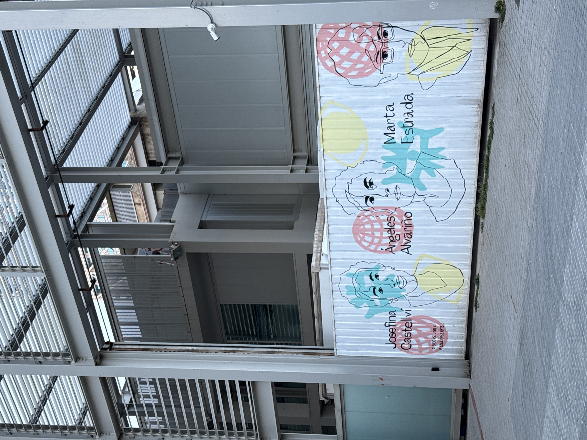 The image shows a multi-story building with a metal framework. Attached to the exterior wall of the building is a banner with portraits of three women: Marta Estrada, Ángeles Alvariño, and Josefina Castellví. The banner features their names alongside stylized line drawings of their faces, each accompanied by geometric shapes in yellow, pink, and light blue. The banner also includes text in a smaller font at the bottom. The ground to the right of the banner is paved with light gray concrete tiles. The scene is potentially located in Barcelona, Spain.