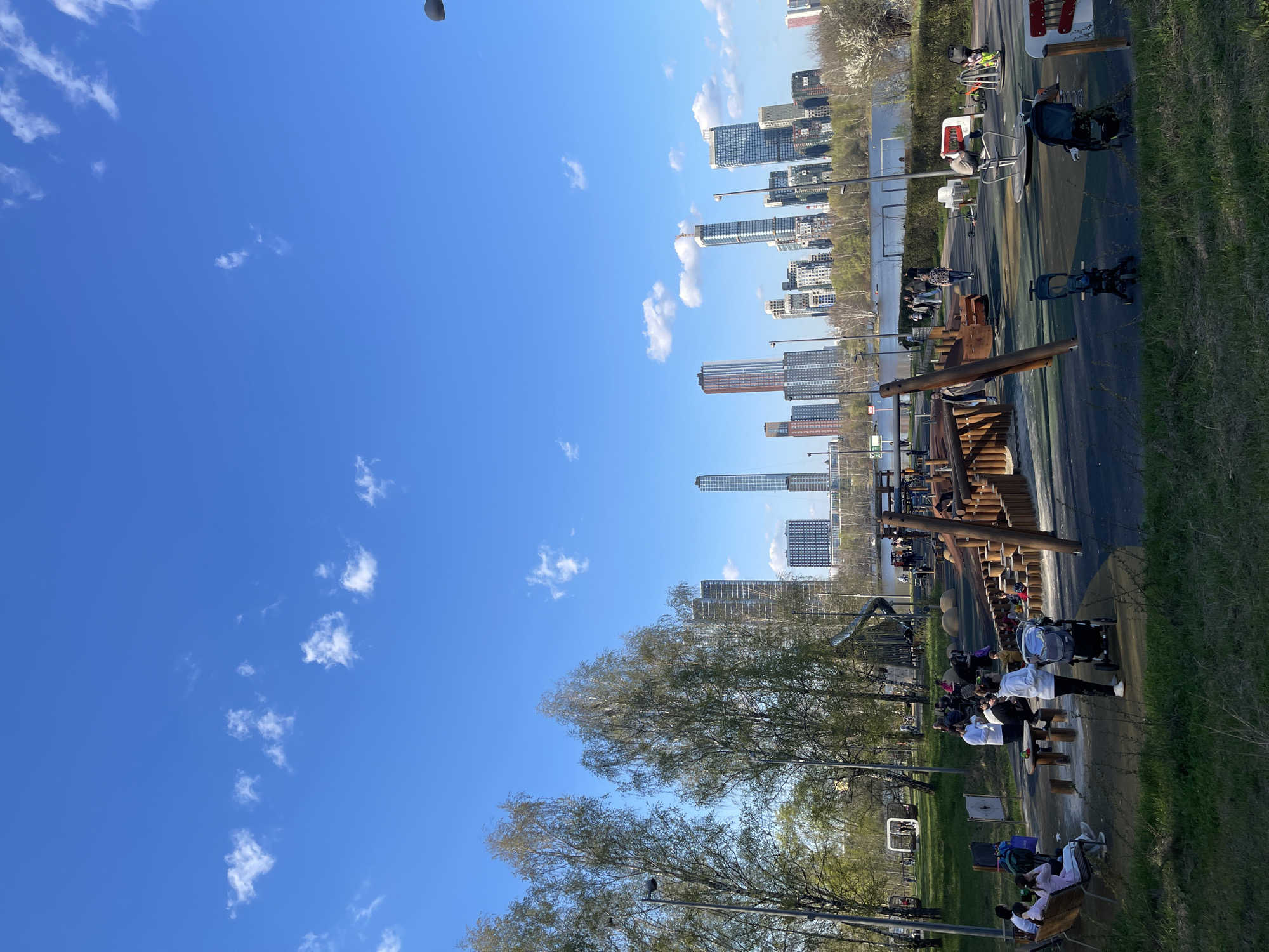 The image shows a view of a city park in Khimki, Russia, during daytime. Tall buildings form the skyline in the background. A playground with wooden structures is visible in the park. Several people, including adults and children, are present. Some are sitting on benches, while others are walking. Strollers are also present. The sky is blue with scattered clouds. Trees with early spring foliage line the park.