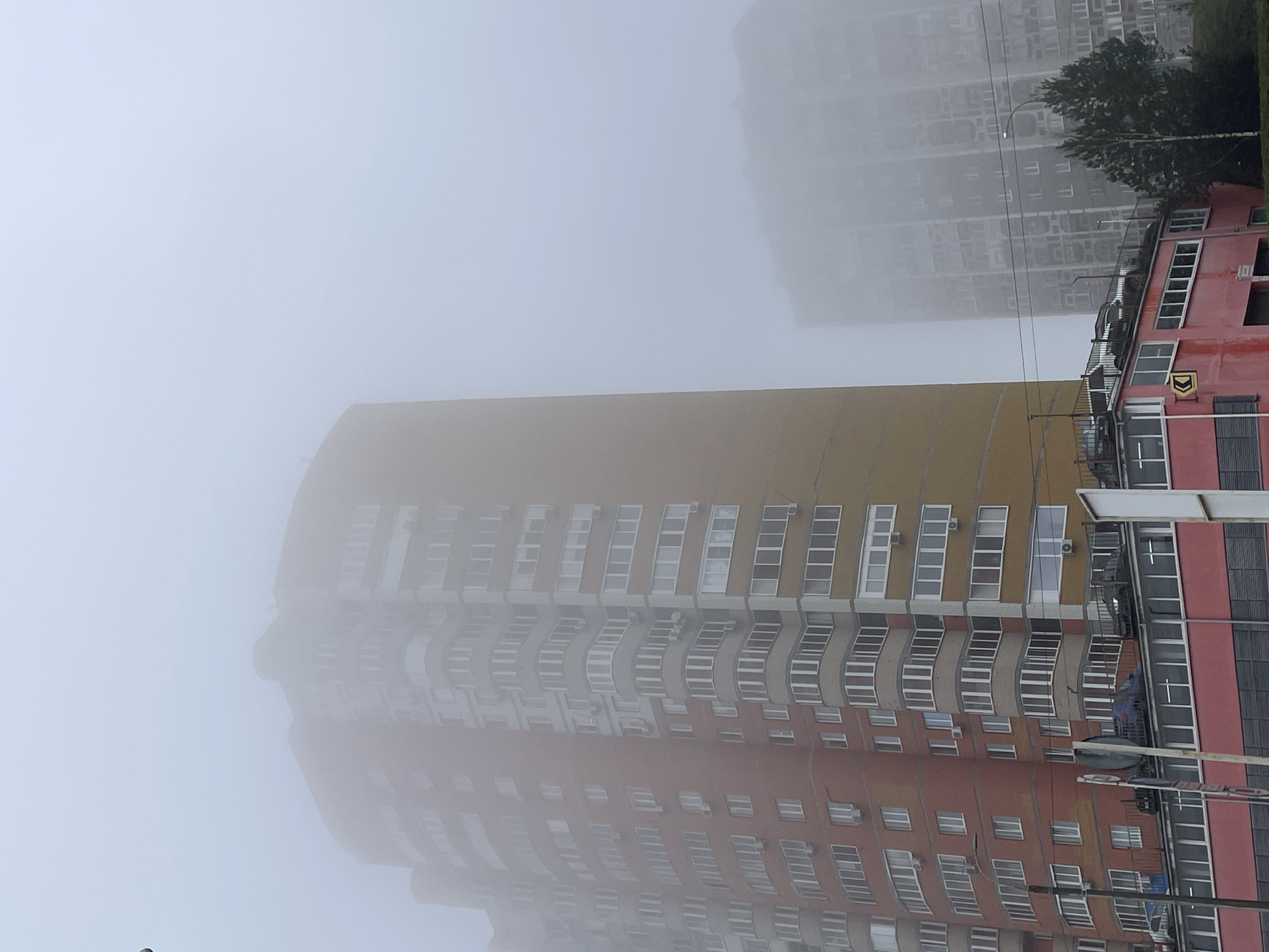 The image depicts a foggy cityscape, likely in Izhevsk, Russia. A tall, multi-story building with a curved facade is partially obscured by fog. The building has a color scheme of yellow and red, with rows of white-framed windows. Another building, with a more conventional rectangular design, is visible in the background, also shrouded in fog. This building seems to have a similar color palette. Overhanging the building on the right are trees and powerlines. The overall impression is of a dense fog obscuring the urban landscape.