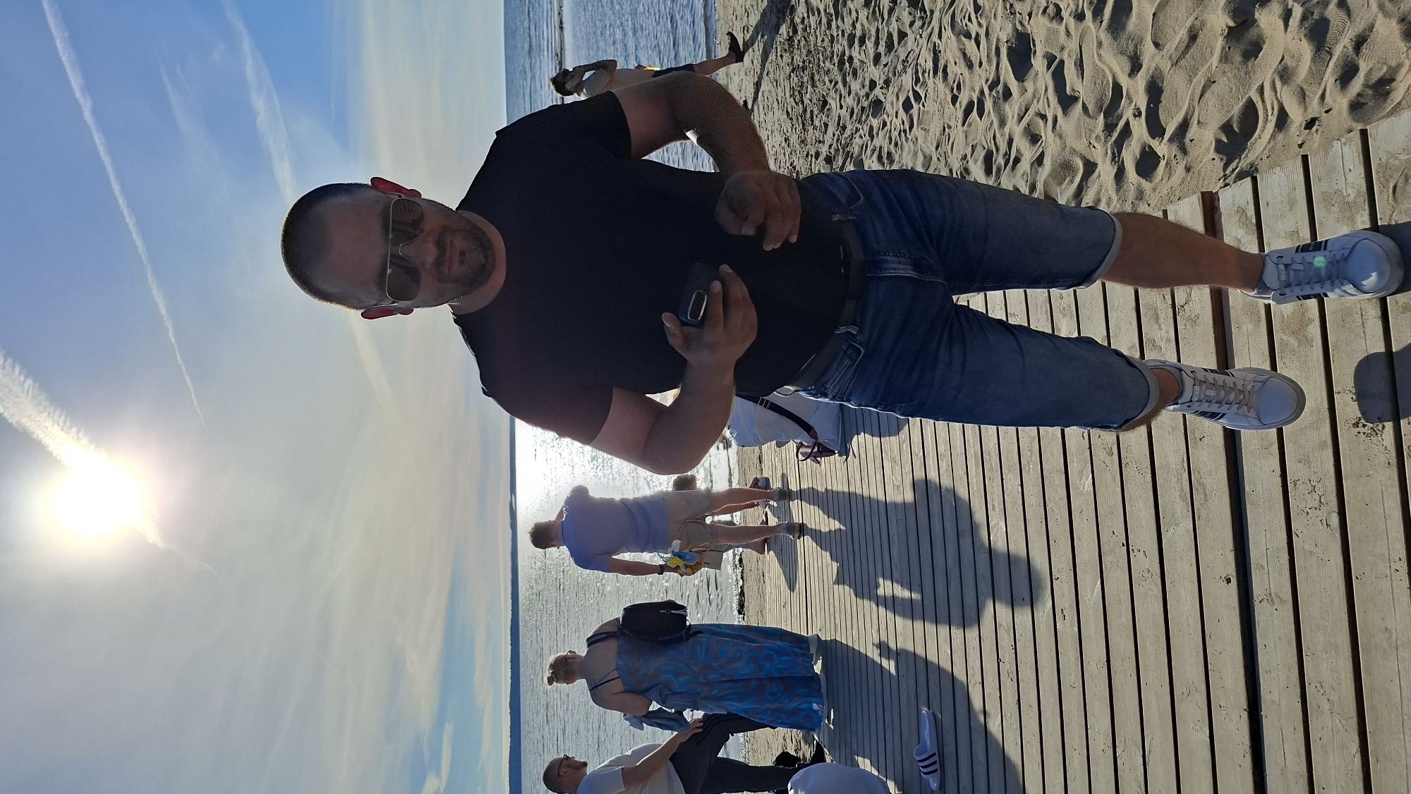 In a sunny outdoor scene at a beach in Tallinn, Estonia, a man is walking on a wooden boardwalk towards the viewer. He is wearing a black t-shirt, blue denim shorts, sunglasses, and white sneakers with black stripes. He holds a black smartphone in his right hand and is looking down towards it. Behind him, several other people are visible on the boardwalk and the beach. To the left, two men and a woman in a blue patterned dress are standing. To the right, a man in a light blue shirt is walking away from the camera, and further back, another person is walking on the sand. The sky is clear blue with a bright sun casting strong shadows. The ocean is visible in the background, reflecting the sunlight. The boardwalk leads to a sandy beach area.