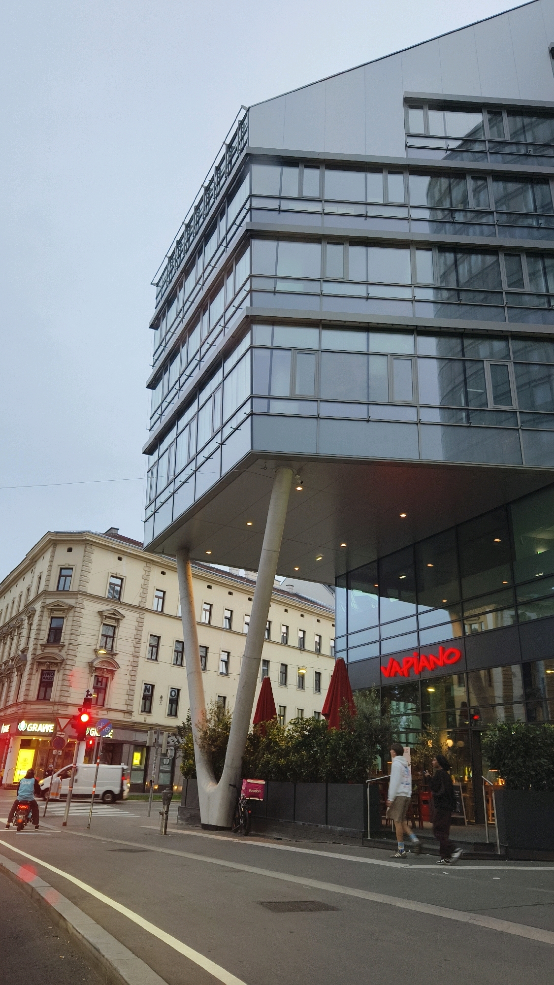 In Vienna, Austria, on a cloudy day, a modern glass-fronted building with a cantilevered section supported by two angled columns stands prominently. The building's facade reflects the overcast sky and surrounding architecture. Below, a red Vapiano restaurant sign is visible. To the left, a traditional cream-colored building with multiple windows and decorative architectural details stands. A street with a pedestrian walkway and a bike lane runs in front of the buildings. A white delivery van is parked near a traffic light which is red. A person on a scooter with a blue backpack is stopped at the traffic light. Two individuals are walking on the sidewalk in front of the Vapiano restaurant. Red umbrellas are visible on an outdoor seating area of the restaurant. A bicycle with a pink food delivery bag is parked near the base of the cantilevered building.