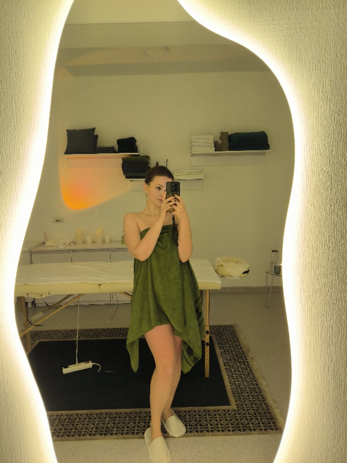 A female individual is depicted taking a mirror selfie in what appears to be a spa or wellness facility located in Vinnytsia, Ukraine. The subject, of fair complexion with light brown hair pulled back, is wrapped in a dark green towel extending from her chest to mid-thigh, and wears light-colored slip-on slippers. She holds a black smartphone with both hands to capture her reflection, partially obscuring her face. The selfie is taken in a large, irregularly shaped mirror featuring integrated warm LED backlighting along its curved edges. The surrounding wall exhibits a textured, light-colored finish.
Reflected in the mirror, the background shows a professional massage table with a white sheet positioned centrally. To the left of the table, a white cabinet or counter holds several white cylindrical objects, possibly candles, beneath an orange ambient light source projecting onto the wall. To the right, white shelving units display neatly folded towels in shades of white and dark green. A small, light-colored side table with a clear container is also visible. The floor is covered by a dark, rectangular rug or mat with a patterned border. A white electrical power strip with a white cord lies on the floor in front of the massage table. The overall lighting is warm and indirect.