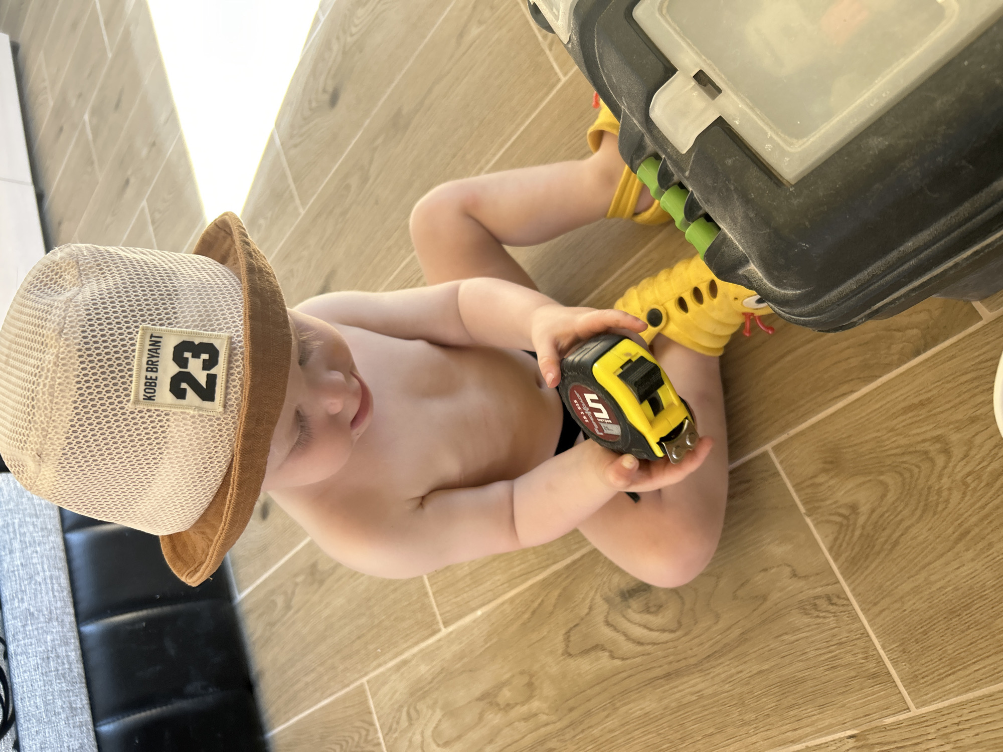 A toddler sits cross-legged on a light brown, wood-patterned tiled floor. The child wears a light brown and mesh hat with a "KOBE BRYANT 23" patch, and yellow Crocs. The child is bare-chested and holds a yellow and black tape measure. Beside the child is a large, black and gray tool box. A portion of a gray sofa with black leather accents is visible in the background. The location is a residence in the city of Chaikovsky, Russia.