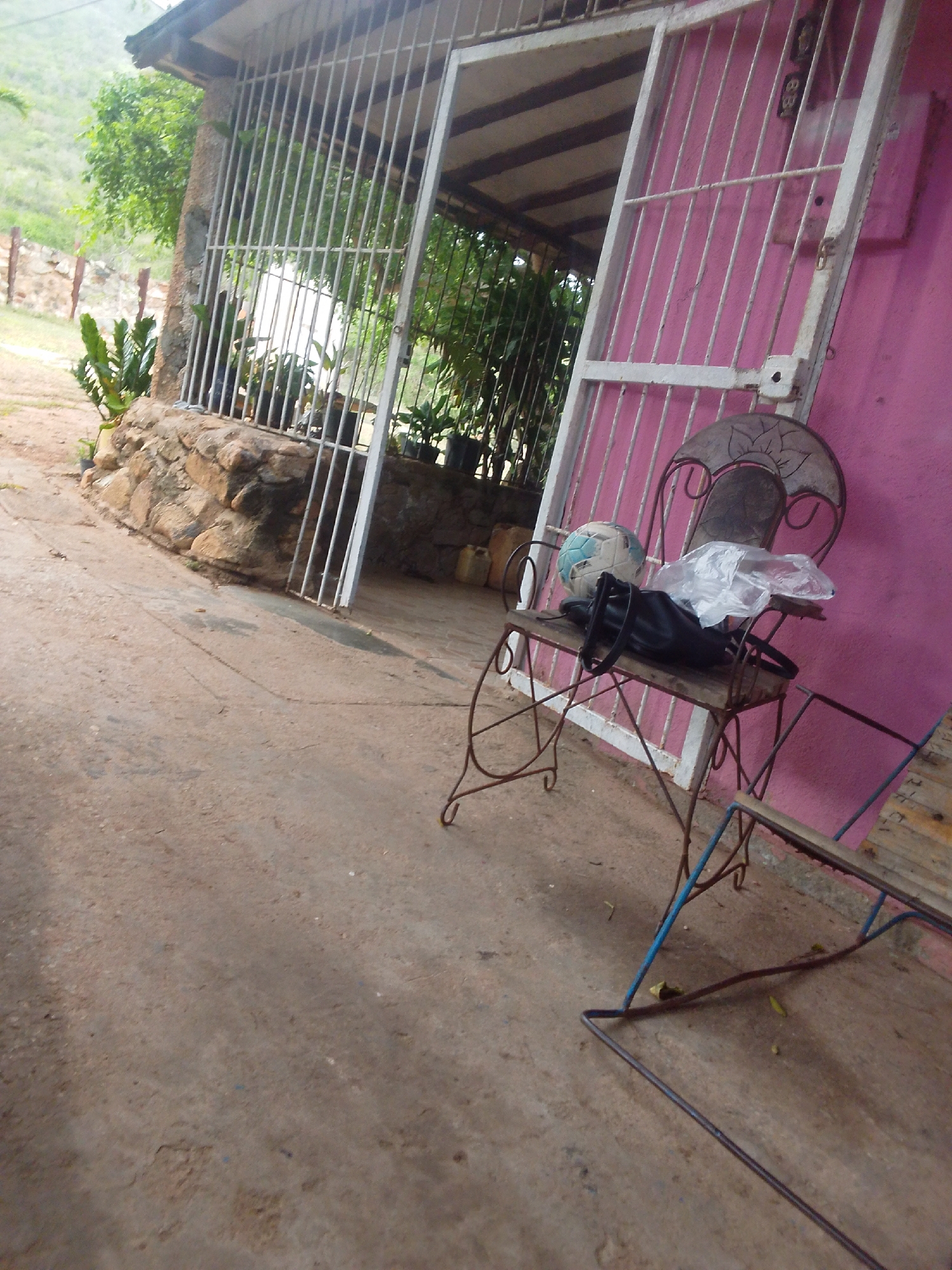 The image shows an outdoor scene in Guayacán, Venezuela, featuring a weathered, pink stucco building with a white metal gate and bars. In front of the building, a rusty, wrought-iron chair sits on a concrete ground. A soccer ball and a black bag with a white plastic bag on top rest on the chair's seat. Behind the chair, more white metal bars and a gate are visible, leading into a shaded area with plants and what appears to be a stone wall. The ground is a light brown concrete, and there are some scattered leaves. In the background, a lush green hillside is visible under an overcast sky.