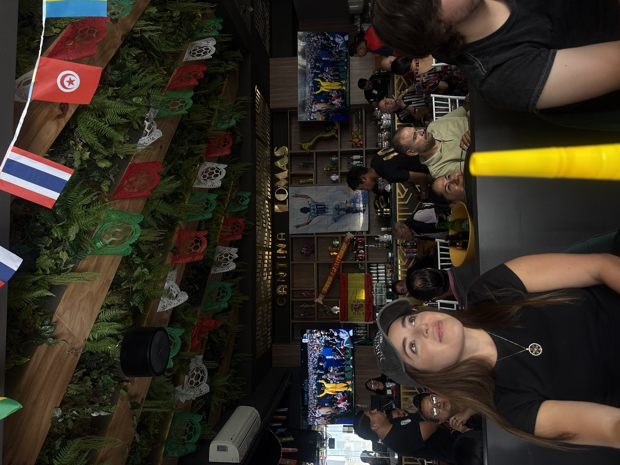 The image depicts the interior of "Cantina Lomas" in San Bernardino Tlaxcalancingo, Mexico. The cantina features a decorative ceiling adorned with lush green foliage and hanging banners, including national flags such as Thailand, Tunisia, and Ukraine, along with traditional Mexican papel picado. Two large television screens are mounted on the wall, displaying sports events. In the foreground, a woman wearing a dark t-shirt and a gray baseball cap is visible, looking upwards. Other patrons are seated around the cantina, some engaging with the screens or at tables. Shelving units behind the bar display bottles, glassware, and decorative items, including a Spanish flag. The overall atmosphere appears to be that of a lively sports bar.