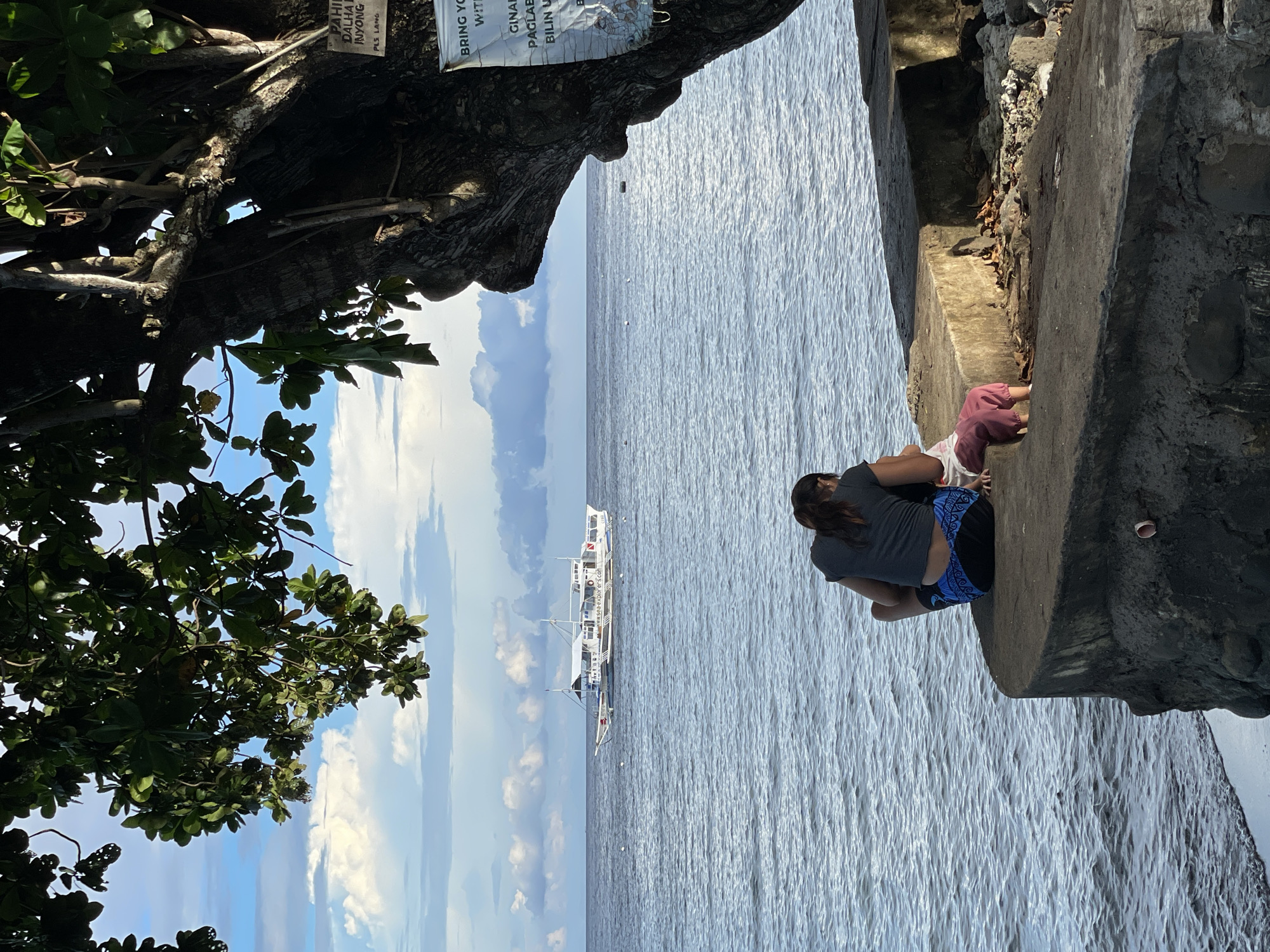 In Dauin, Philippines, two individuals, likely a mother and child, are seated on a concrete embankment overlooking the sea. The woman, wearing a gray t-shirt and patterned shorts, is in a seated position, leaning slightly forward. The child, dressed in a pink garment, is in front of her, also seated. Their backs are to the camera, suggesting a moment of quiet observation.
The sea, a calm expanse of blue water, stretches towards the horizon where a white passenger boat with multiple decks is visible. The boat appears to be anchored or moving slowly. The sky above is a mix of blue and white clouds, creating a gentle contrast with the water.
To the right of the frame, a large tree with dark, textured bark partially obscures the view. Its branches, laden with green leaves, hang over the scene, casting shadows on the embankment. A weathered sign is attached to the tree trunk. The embankment itself is made of rough concrete, with visible rocks and some debris scattered on its surface. A small white plastic object, possibly a cap or lid, lies on the concrete near the edge. The overall scene evokes a peaceful, seaside ambiance, possibly a local park or public access area along the coast of Dauin.