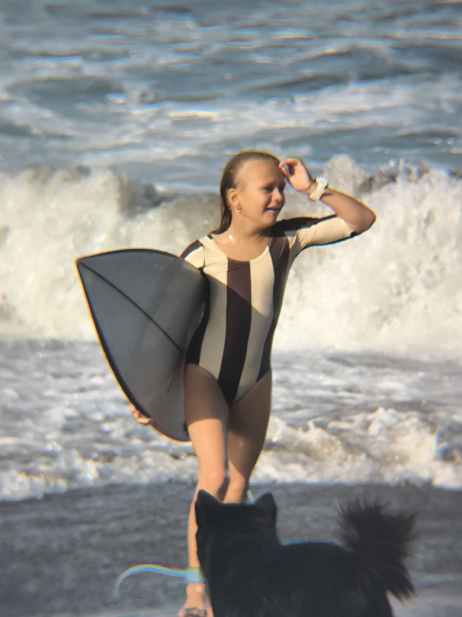 A young girl in a striped swimsuit stands at the shoreline holding a surfboard, shielding her eyes with her right hand. A black dog stands near her legs. Behind them are waves breaking on the shore. The location is likely in North Kuta, Indonesia.