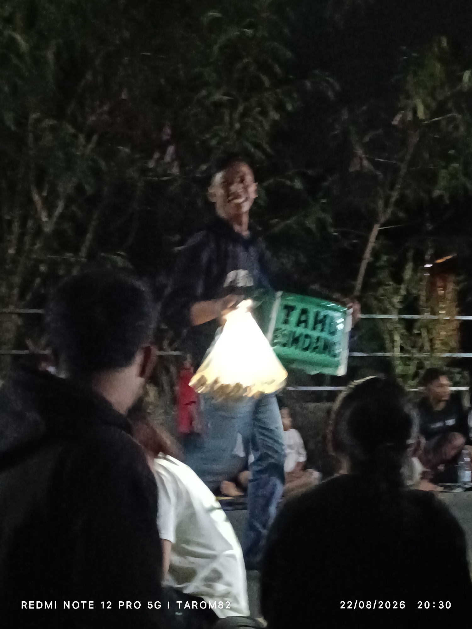 The image, captured at night, depicts an outdoor gathering in Mundusewu, Indonesia, likely in a semi-rural setting given the presence of dense foliage in the background. In the center, a young man, wearing a dark jacket and jeans, is holding a glowing plastic bag in his left hand and a green sign with white lettering in his right. The sign appears to read "TAHU ESMONDANG," with "ESMONDANG" partially obscured. The man is smiling and looking towards the camera.
In the foreground, the backs of several people are visible, suggesting they are observing the central figure or the event. One individual on the far left wears a black hoodie. Another person to the right is in a white t-shirt. The overall lighting is dim, with the bright plastic bag and the sign serving as primary light sources for the central subject. The presence of a date and time stamp in the lower right corner, "22/08/2026 20:30," and a device identifier, "REDMI NOTE 12 PRO 5G | TAROM82," suggest the image was taken with a smartphone. The environment appears to be somewhat informal, with elements of a community gathering or performance.