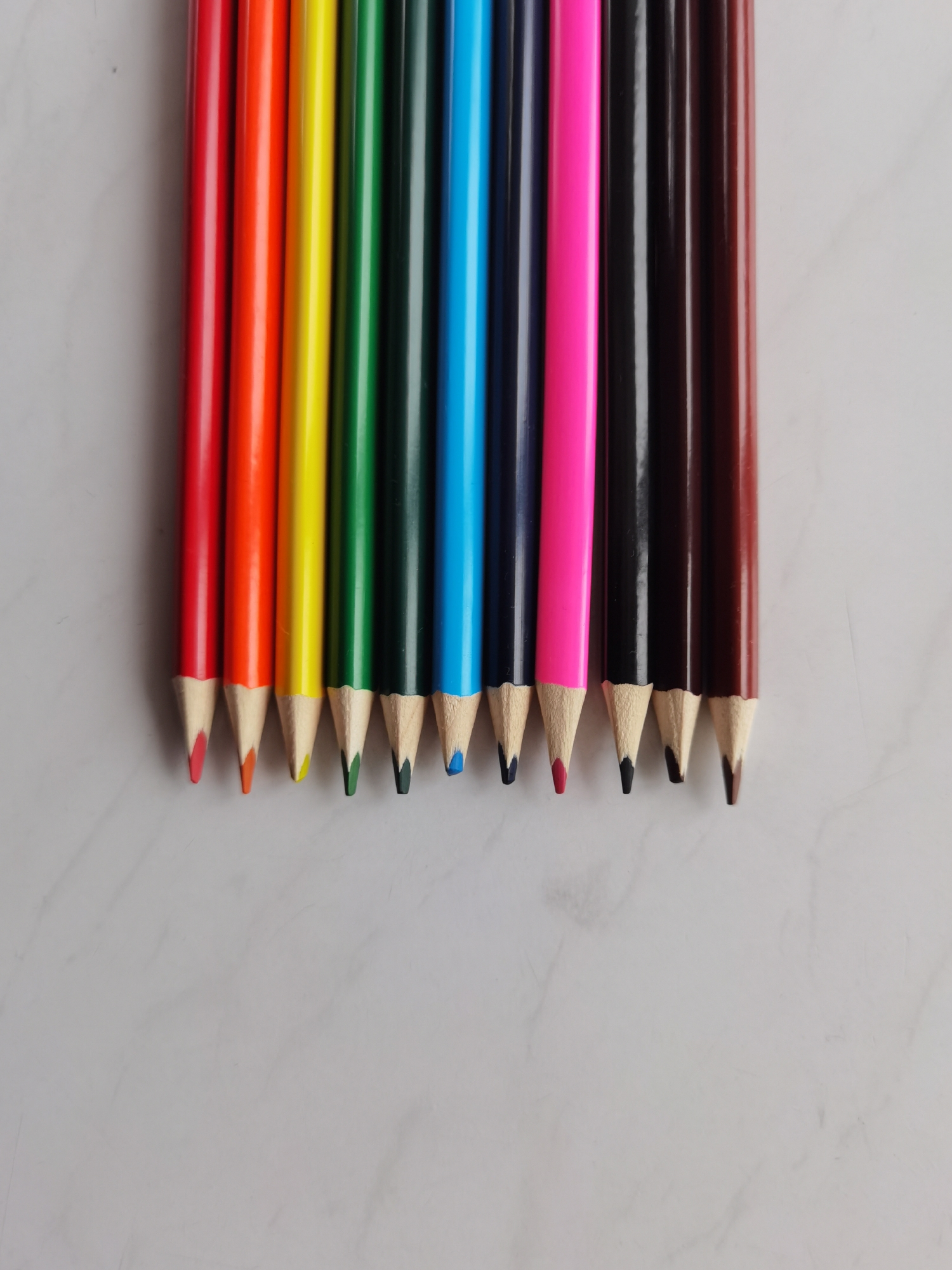 The image shows a set of ten colored pencils arranged horizontally on a light gray, marble-patterned surface. The pencils are aligned with their points facing downward. From left to right, the colors are red, orange, yellow, green, dark green, blue, navy blue, pink, black, and brown. The pencils are new or recently sharpened, with the colored lead tips exposed. There are no indications regarding the location in Perm, Russia.