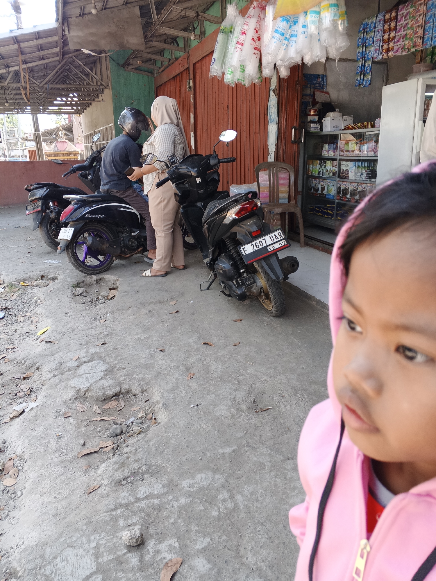 In Sukabumi, Indonesia, outside a small storefront with a corrugated metal roof and orange wooden doors, two individuals stand beside motorcycles on a cracked and uneven concrete surface littered with dried leaves. A male figure wearing a dark shirt and helmet is seated on a black scooter. A female figure in a plaid shirt and a light-colored hijab stands next to him, holding a smartphone. Another black scooter is parked in front of them. The storefront displays various items, including plastic bags of disposable cups hanging from the ceiling and shelves stocked with packaged goods inside a glass-fronted cabinet. A white refrigerator is visible to the right. In the foreground, the blurred image of a young child wearing a pink hooded jacket suggests a close-up perspective, possibly indicating the photographer. The scene appears to be a casual stop at a local convenience store or warung.