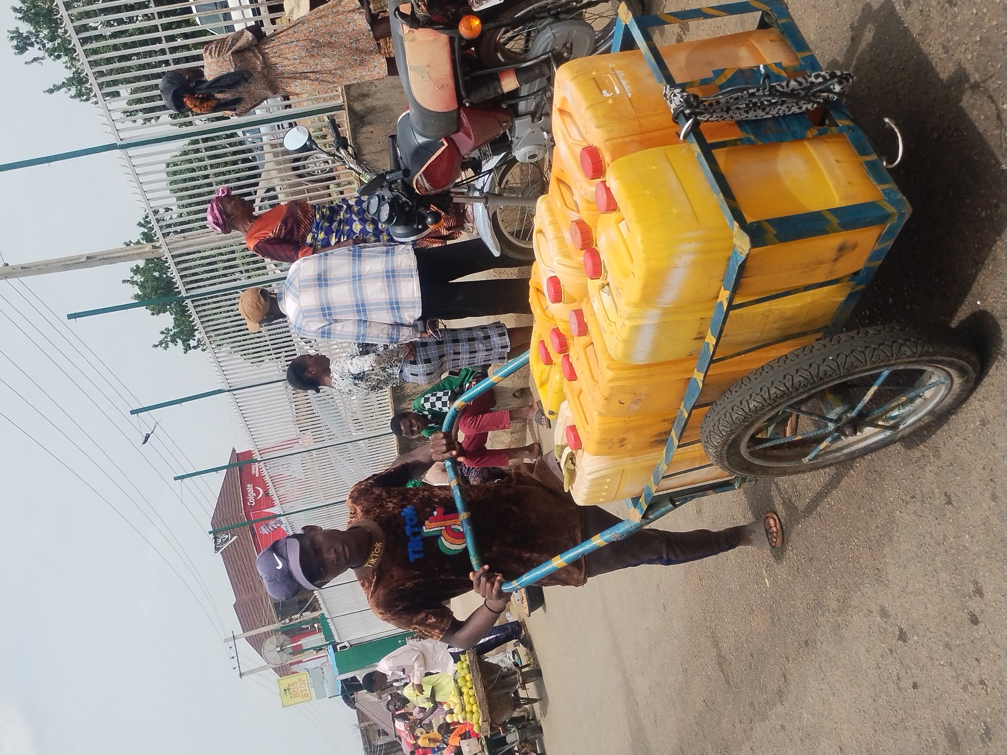 A man is pulling a hand-drawn cart laden with yellow jerrycans down a street in Jos, Nigeria. The man is wearing a brown shirt with "TIKTOK" on the collar and a purple baseball cap. The cart is blue and yellow, and the jerrycans have red caps. Other people are visible in the background, including some standing near a motorcycle. A sign for a "Bus Stop" and a Colgate advertisement are also visible in the background.