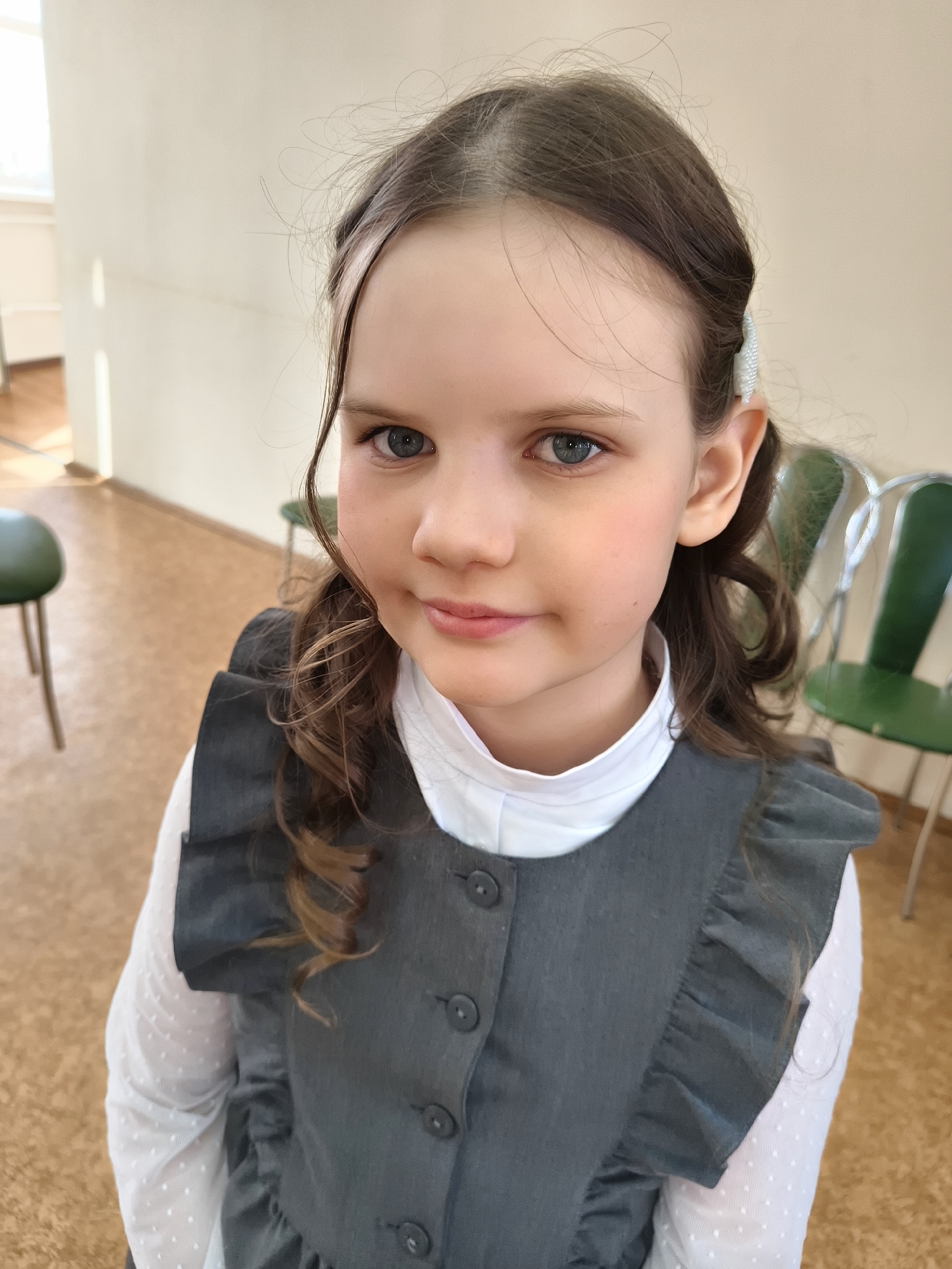 A young girl is captured in a close-up, eye-level portrait shot, looking directly at the camera with a subtle, pleasant smile. She has light brown hair, with some strands loosely framing her face, and striking blue eyes. A white, beaded hair clip is visible on the right side of her head. She is dressed in a white long-sleeved shirt with a delicate polka dot pattern, layered under a grey jumper dress or pinafore featuring ruffled shoulders and a row of dark buttons down the front.
The setting appears to be indoors, likely a school or institutional building, characterized by plain, light-colored walls and a light brown, possibly cork, floor. Bright natural light illuminates the scene, suggesting daytime. In the blurry background, parts of two green chairs with metallic frames are visible, consistent with a classroom or public space. The girl's expression conveys a sense of calm and mild curiosity. No text is visible in the image.