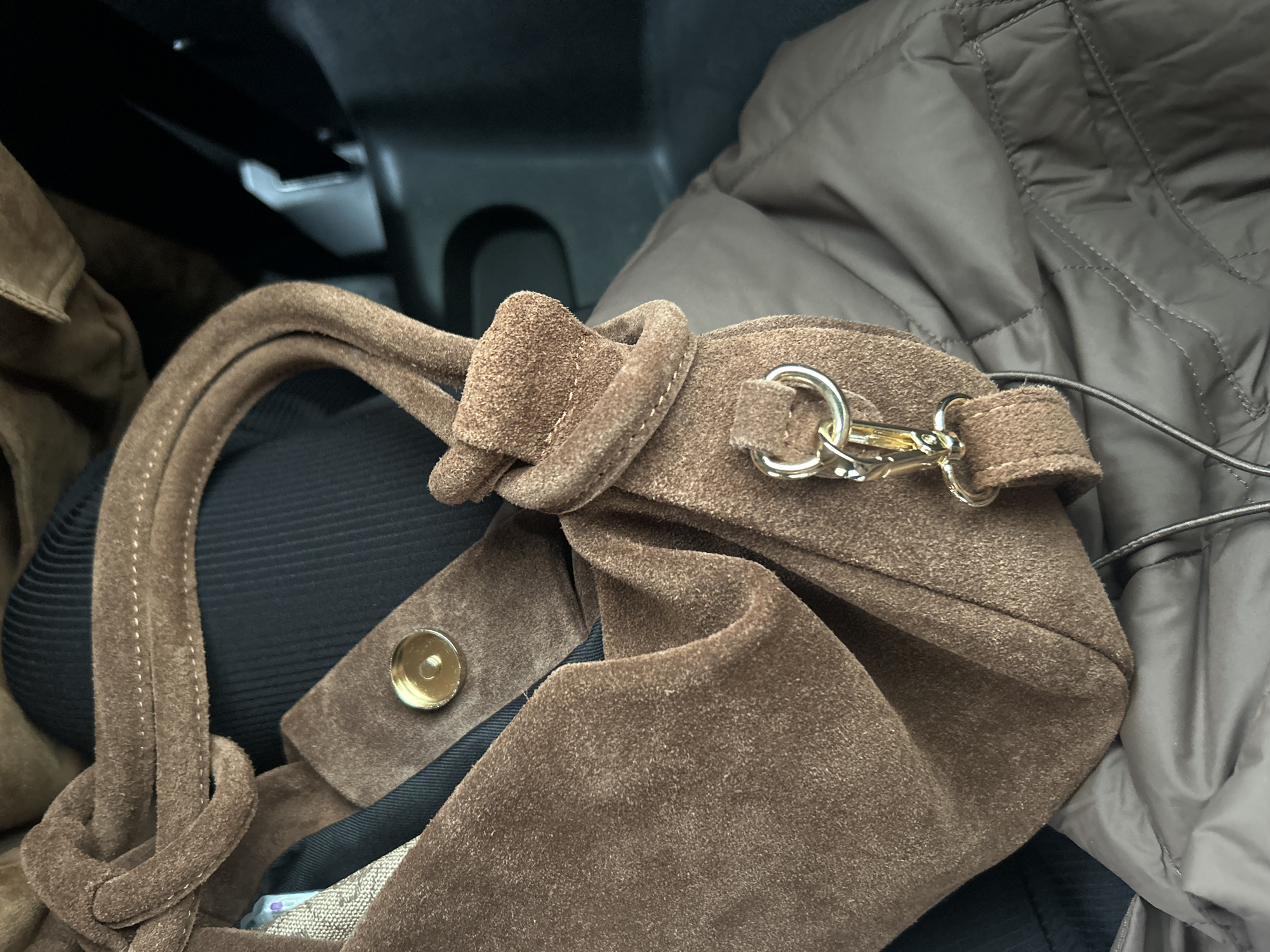 The image is a close-up of a brown suede handbag with gold hardware, resting on a brown puffer jacket. The handbag has a knotted strap and a detachable strap with a clip. A gold snap closure is visible on the bag. The background appears to be the interior of a car, with dark gray plastic elements and the seat. The lighting suggests an indoor environment or a car interior. There is no discernible indication of the location being the city of Chaikovsky, Russia.