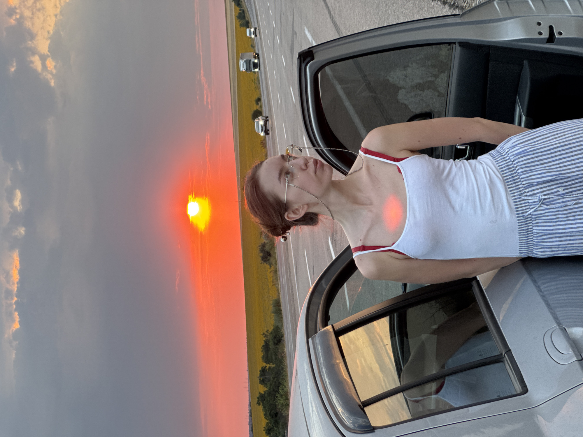 A young woman with auburn hair styled in a bun and wearing a white tank top and blue and white striped pants stands next to a silver car with its doors open. She has on glasses with a chain attached. The car is parked on the side of a road with traffic visible in the background, including trucks and cars. The scene is set against a vibrant sunset with a bright orange sun and dramatic clouds. A field of yellow flowers stretches across the horizon behind the road. The location is identified as Yelyzavetivka, Ukraine.