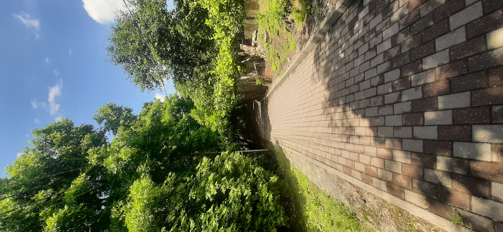 An outdoor perspective shot shows a pathway made of reddish-brown and gray interlocking bricks stretching into the distance. The path is bordered on the left by a gravel area and then lush green grass, followed by dense foliage of trees with vibrant green leaves. On the right, the path is bordered by a concrete curb and then a dirt embankment covered in green vegetation. A low, weathered fence, partially obscured by the greenery, runs along the right side of the path further into the distance. Overhead, the sky is a clear blue with scattered white clouds. The scene appears to be set in a park or a residential area with abundant natural vegetation, possibly in Yessentukskaya, Russia, given the context. No people are visible in the image.