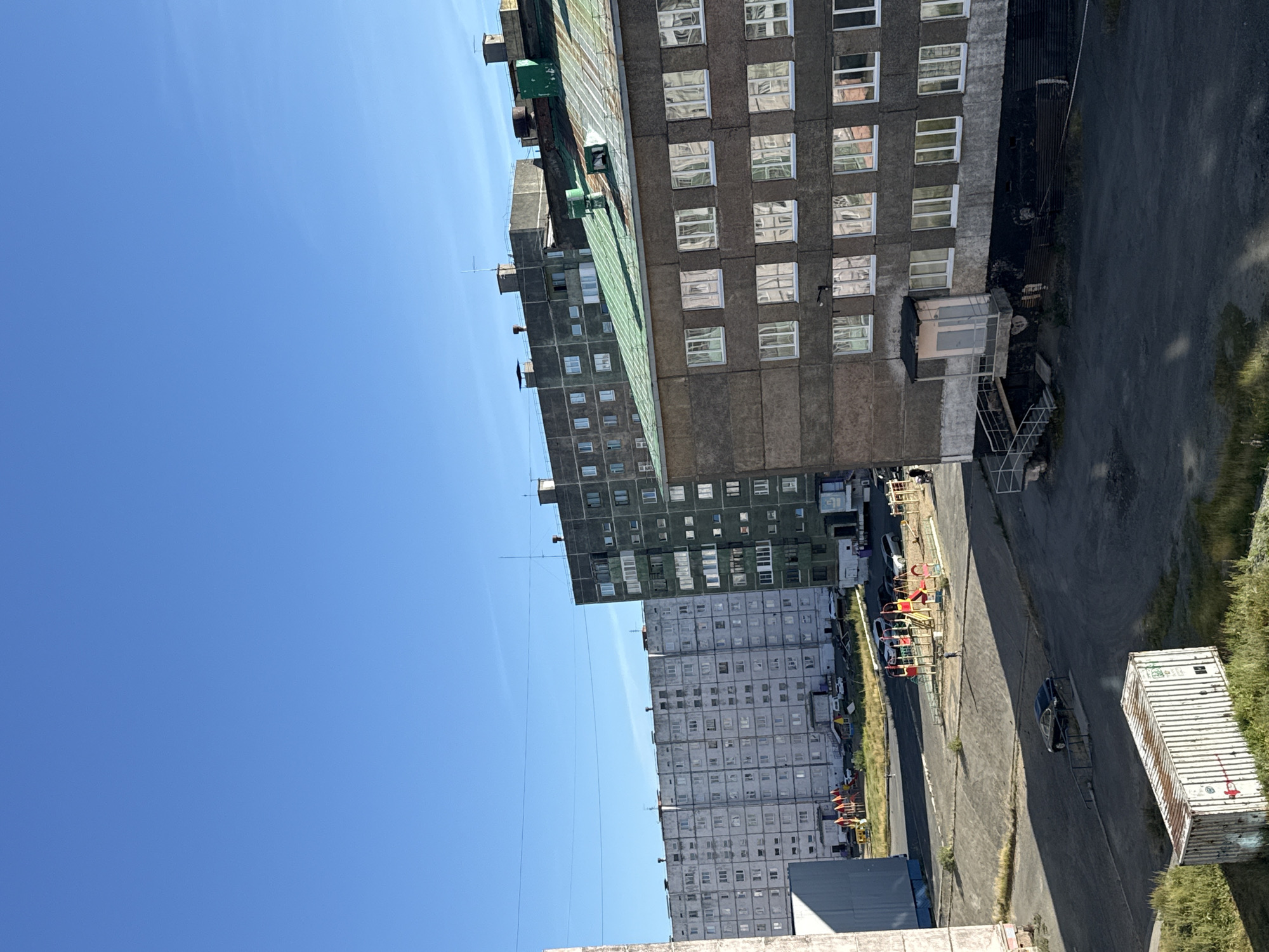 The image shows a view of a residential area in Norilsk, Russia, on a clear day with a bright blue sky. In the foreground, a concrete building with a green, corrugated metal roof is visible, featuring multiple windows reflecting the sky and surroundings. To the left of this building, another concrete structure partially obscures the view. Further in the background, a long, multi-story apartment building with a light gray facade and numerous windows stretches across the scene. To its right, another dark gray apartment building stands, also with many windows. A playground with colorful equipment, including slides and swings, is situated between the buildings. Several cars are parked in the area, and a white shipping container rests on the ground in the lower left portion of the image, surrounded by overgrown grass. The overall environment suggests a northern urban landscape with functional, utilitarian architecture.