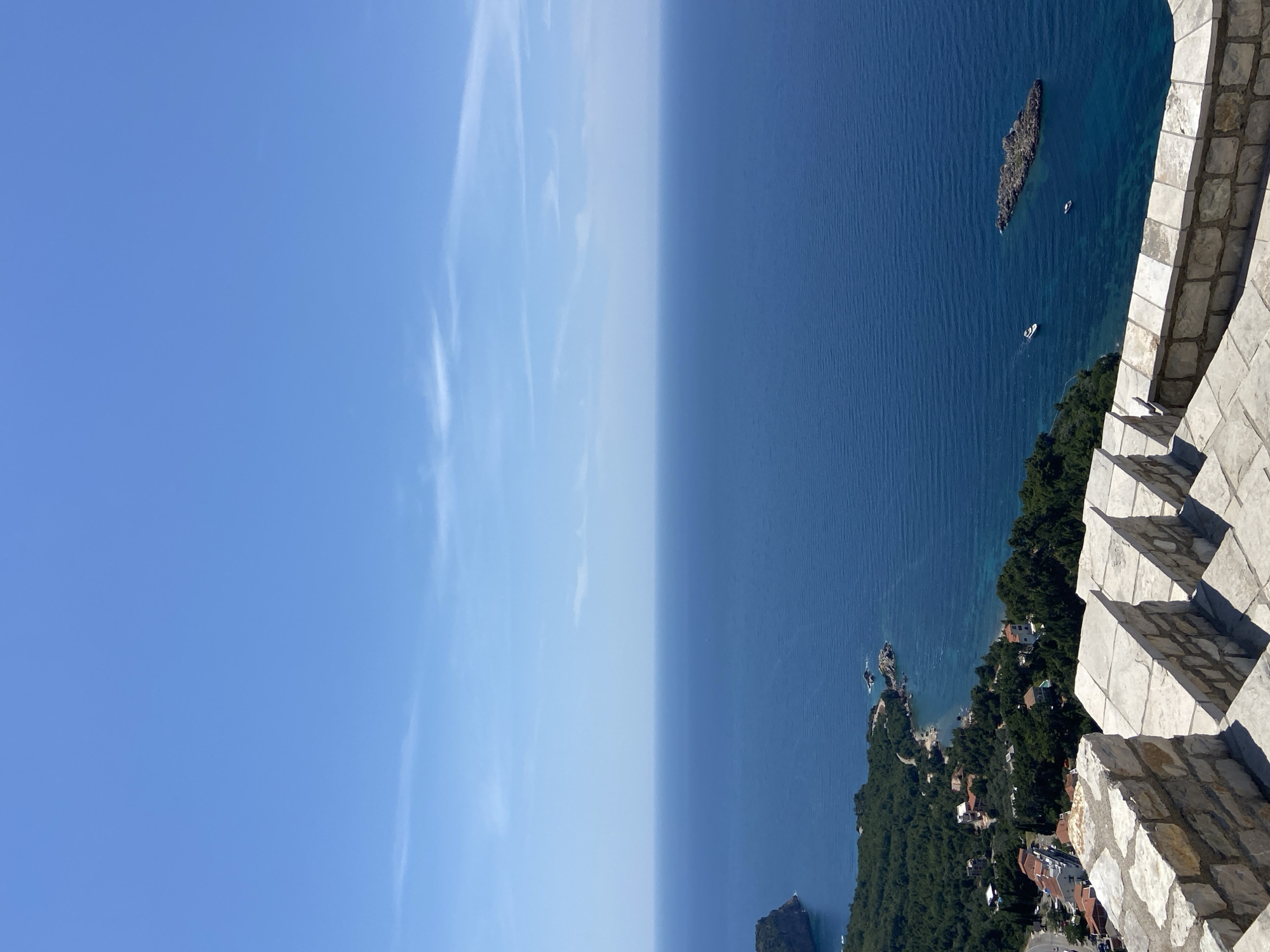 A high-angle, vertical shot captures a vast expanse of the deep blue Adriatic Sea meeting a clear, bright blue sky, with only a few wispy clouds visible near the horizon. In the foreground, a stone staircase with a rough-hewn, textured railing made of light-colored, irregular stones leads the eye down towards the scene.
The middle ground reveals a picturesque coastal landscape, characteristic of Sveti Stefan, Montenegro. Lush green trees densely cover the hillside, interspersed with terracotta-roofed buildings, suggesting a residential area nestled along the coast. A winding road is visible, hinting at accessibility. The coastline is rugged, with rocky outcrops and small coves bordering the water.
Further out in the sea, two small boats, one a white yacht and the other a smaller vessel, are visible. To the right of the boats, a dark, jagged rock formation juts out of the water, a prominent feature in the seascape. The overall impression is one of a serene, sunny day with a stunning view of the Montenegrin coast.