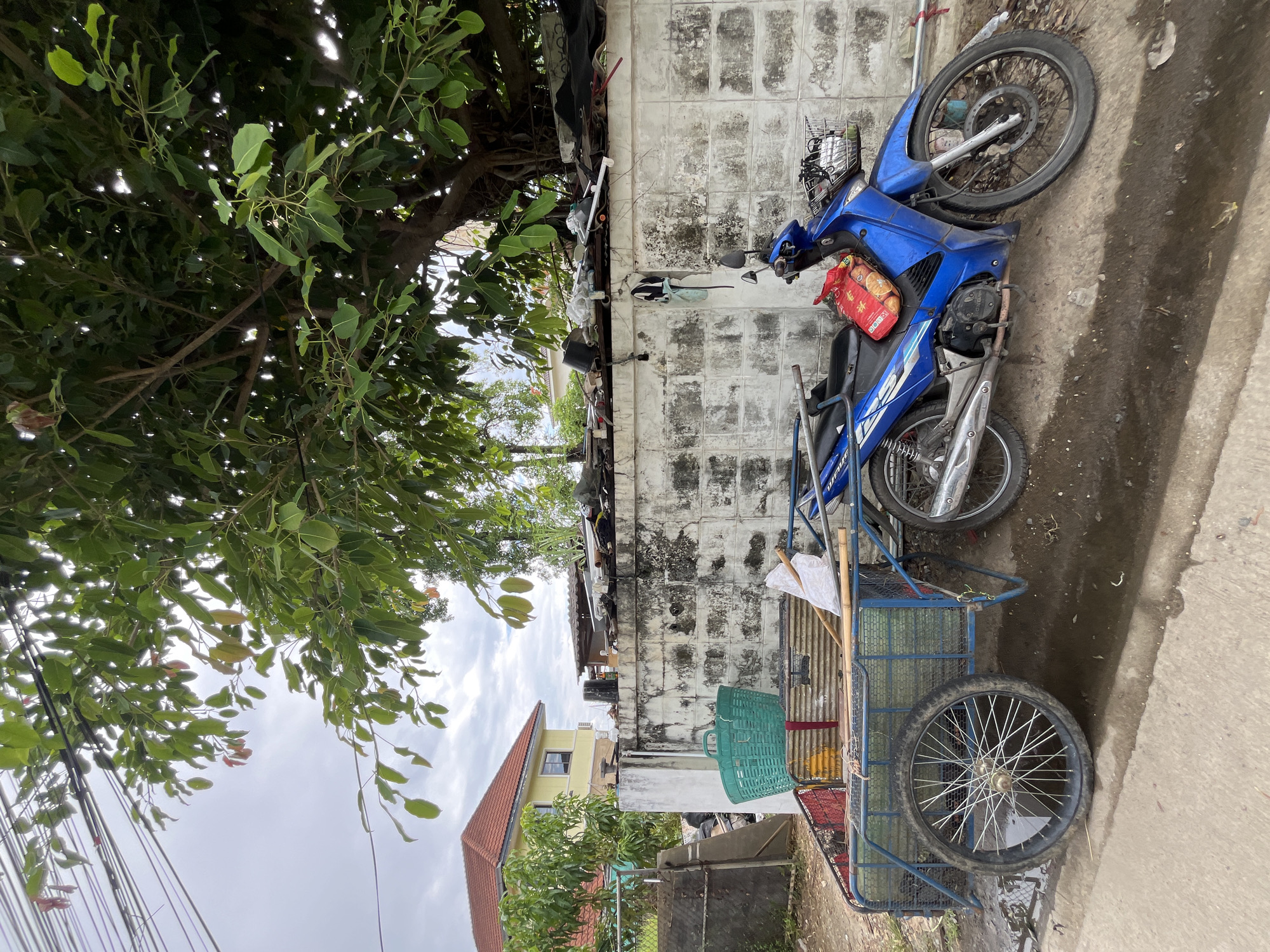 In the city of Ban Kanchanok 23, Thailand, a blue motorcycle is parked next to a two-wheeled cart. The cart is filled with items, including a green basket, bamboo poles, and a white bag. The motorcycle has a basket on the front containing a red bag of goods. Behind the cart and motorcycle is a rough, white cinder block wall, topped with debris and vegetation. A large tree with dense foliage overhangs the scene from the right. In the background, a yellow house with a red roof is visible, along with a chain-link fence. The ground is a mix of dirt, concrete, and puddles of water, suggesting recent rain. Overhead, electrical wires are strung between poles. The overall scene depicts a cluttered, outdoor environment, possibly a residential or commercial area.