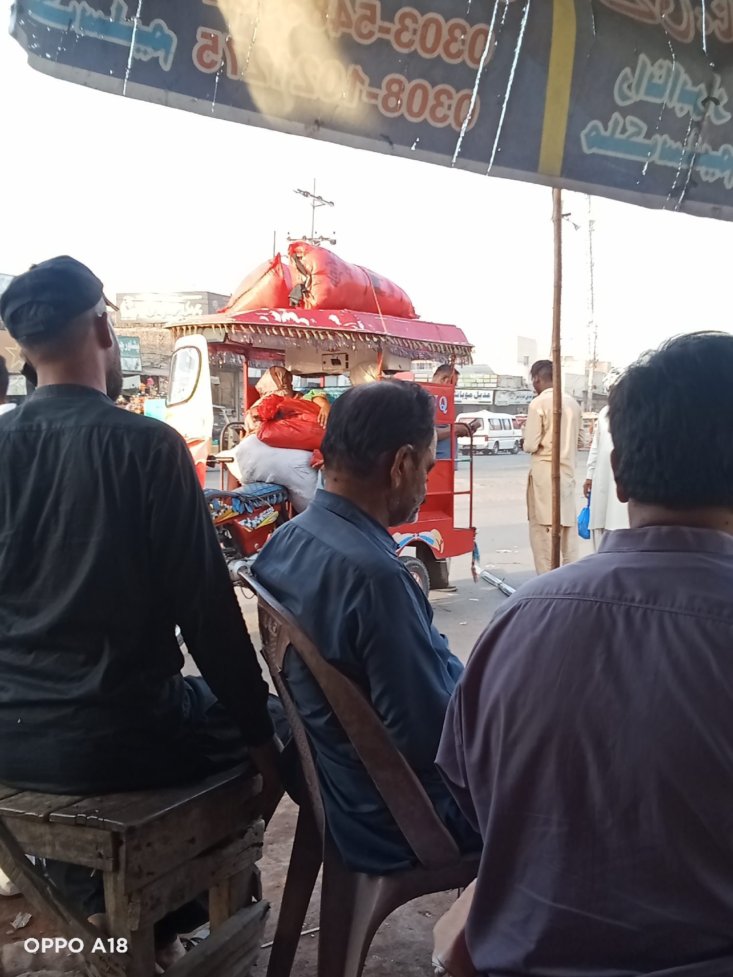 Here's a forensic description of the image:
The image shows three men seated under an awning in a city of Unknown, Unknown. The man on the left is wearing a black shirt and cap, sitting on a weathered wooden stool. The man in the center is wearing a dark blue shirt and sitting on a plastic chair, while the man on the right is wearing a gray shirt.
In the background, a red motorized tricycle loaded with white and red bags is parked on the street. A man can be seen on the tricycle. There are other people walking in the background, along with a white van.