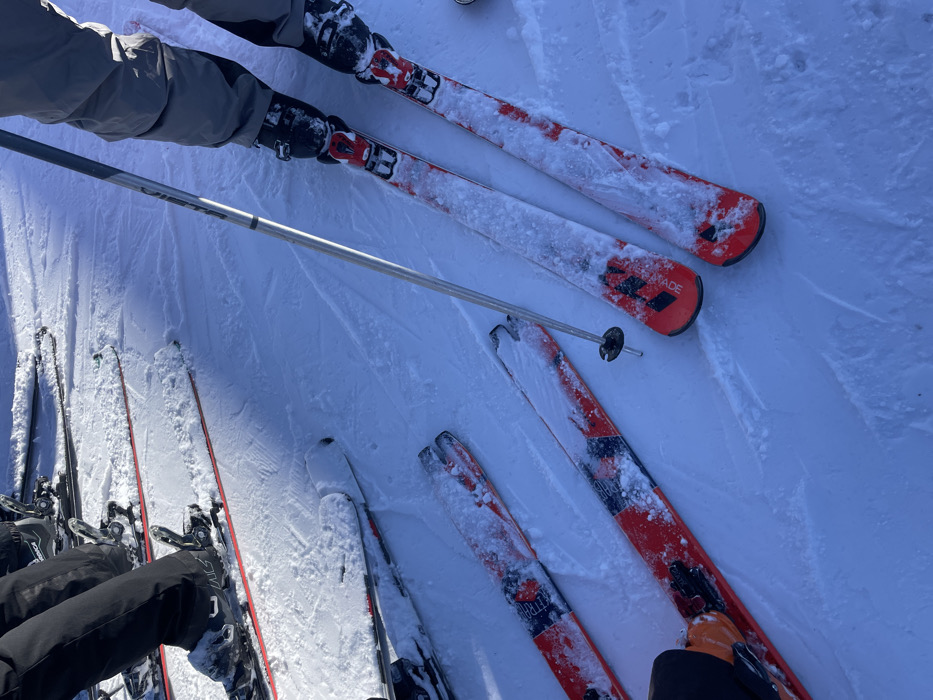 The image provides an overhead view of skiers' legs and equipment on a bright, snow-covered ski slope in Séez, France. At least three individuals are depicted through their ski gear. In the upper part of the frame, a person wearing grey ski pants and black ski boots is standing with their red and black skis, which are partially covered in powdery snow. A silver ski pole extends diagonally from this skier towards the lower right.
Below, multiple pairs of skis are visible resting in the snow. One pair of red and black skis, with "ARMADA" clearly visible on its surface, is prominent in the mid-ground. Another pair of red, black, and white skis, showing fragmented text like "BLITRA" and "900S", is positioned towards the lower right, connected to orange and black ski boots. A third set of black-clad legs and skis can be seen in the lower-left corner.
The scene suggests a moment of rest or preparation during a ski outing. The snow is bright white and textured, with some areas appearing freshly fallen or undisturbed, while others show tracks and compacted surfaces. The strong shadows cast by the equipment indicate bright daylight and clear weather. The skis and boots are dusted with snow, highlighting the cold, wintry conditions of the mountain environment.