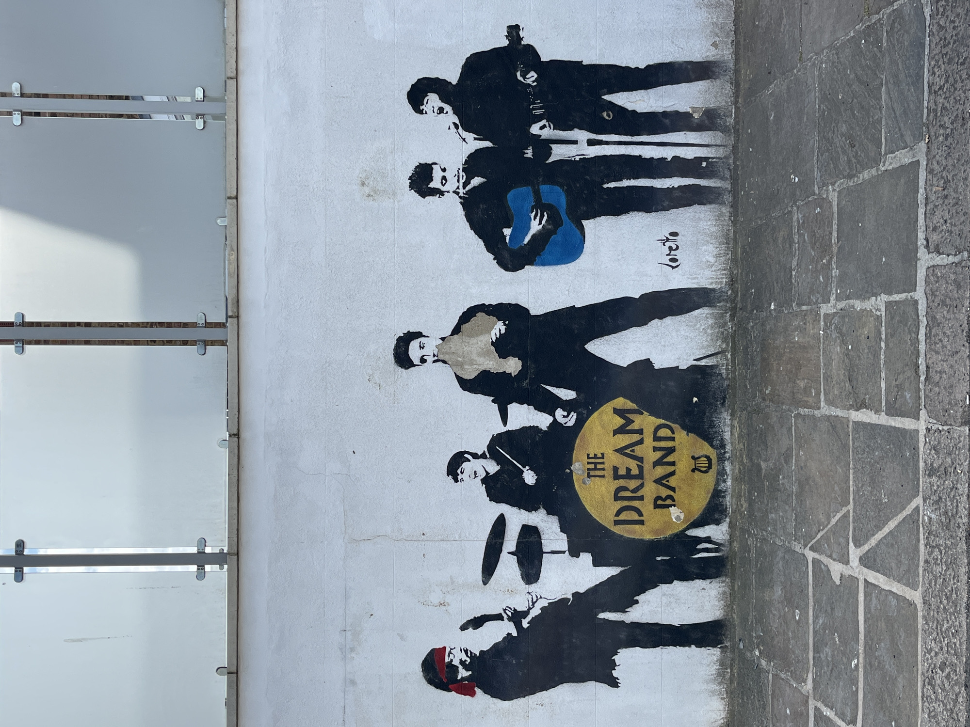 A vertical, medium-shot image depicts a wall with graffiti art of a musical band, adjacent to a glass barrier and a stone-paved surface. The wall is white with black stenciled figures of musicians. From top to bottom: two guitarists, one with a blue guitar, followed by a keyboardist, a drummer with a golden drumhead that reads "THE DREAM BAND," and finally a bassist wearing a red bandana. The graffiti art extends downward to the rough edge of a stone-paved surface. To the left of the graffiti, there is a glass barrier consisting of multiple rectangular glass panels, held in place by metal brackets. The photo was taken in Unknown, Unknown.