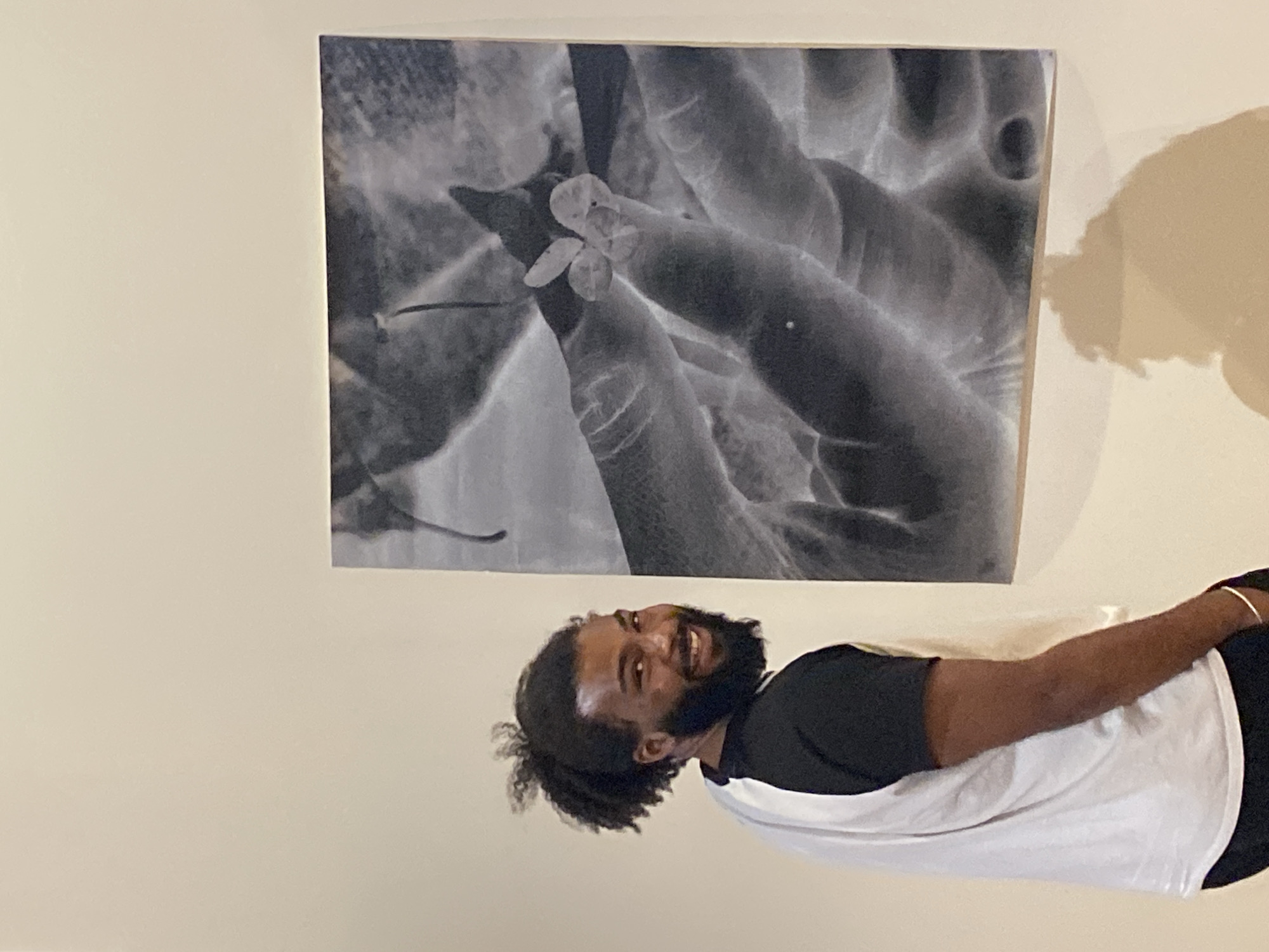 An indoor, medium shot captures a smiling man standing next to a large monochrome artwork displayed on a light-colored wall. The man, with dark skin, dark curly hair styled back, and a full beard and mustache, is seen from the chest up, oriented slightly to the left. He wears a black short-sleeved shirt with a white sleeveless top or undershirt layered over it, and a subtle gold-colored bracelet is visible on his left wrist. His expression is one of broad, joyful smiles, looking directly towards the viewer.
To his upper left, a large, rectangular, black and white artwork hangs on the wall. The artwork depicts what appears to be an inverted or X-ray-like image of hands gently holding or interacting with small, translucent, berry-like or floral elements. The background of the print is soft and blurred, suggesting other textures or plant forms. The wall behind the man and the artwork is a uniform, neutral cream or off-white color. A soft shadow is cast on the wall to the right of the artwork, and a larger, indistinct shadow falls on the right side of the frame, possibly from the man or another light source. No discernible text is visible in the image. The setting is indoors, possibly a gallery, home, or exhibition space, with the given context being Paris, France.