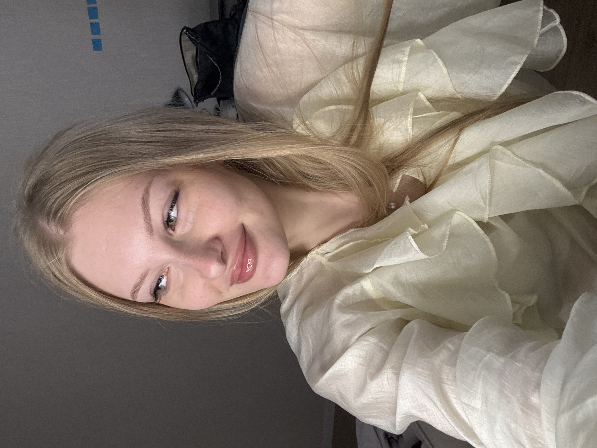 A young woman with blonde hair and fair skin is looking to her left with a slight smile. She is wearing a light yellow ruffled shirt. Her makeup includes mascara, eyeliner, and lip gloss. Her hair is long and straight, with some strands falling over her shoulder. In the background, a dark bag is visible on a surface. The wall behind her has a few blue square decals. The lighting is dim, creating shadows. The overall scene appears to be indoors, possibly a bedroom, and there are no discernible landmarks or architectural features that would indicate a specific location in the city of Perm, Russia.