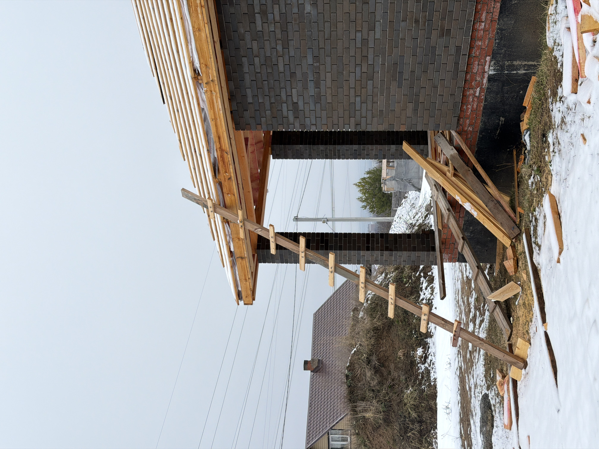 The image captures a construction site in what is presumed to be Chaikovsky, Russia, based on the prompt. A partially constructed brick building stands alongside a crude wooden ladder, leaning against the structure. Wood and snow are seen around the base. In the distance, parts of other buildings and trees are visible under a light sky.