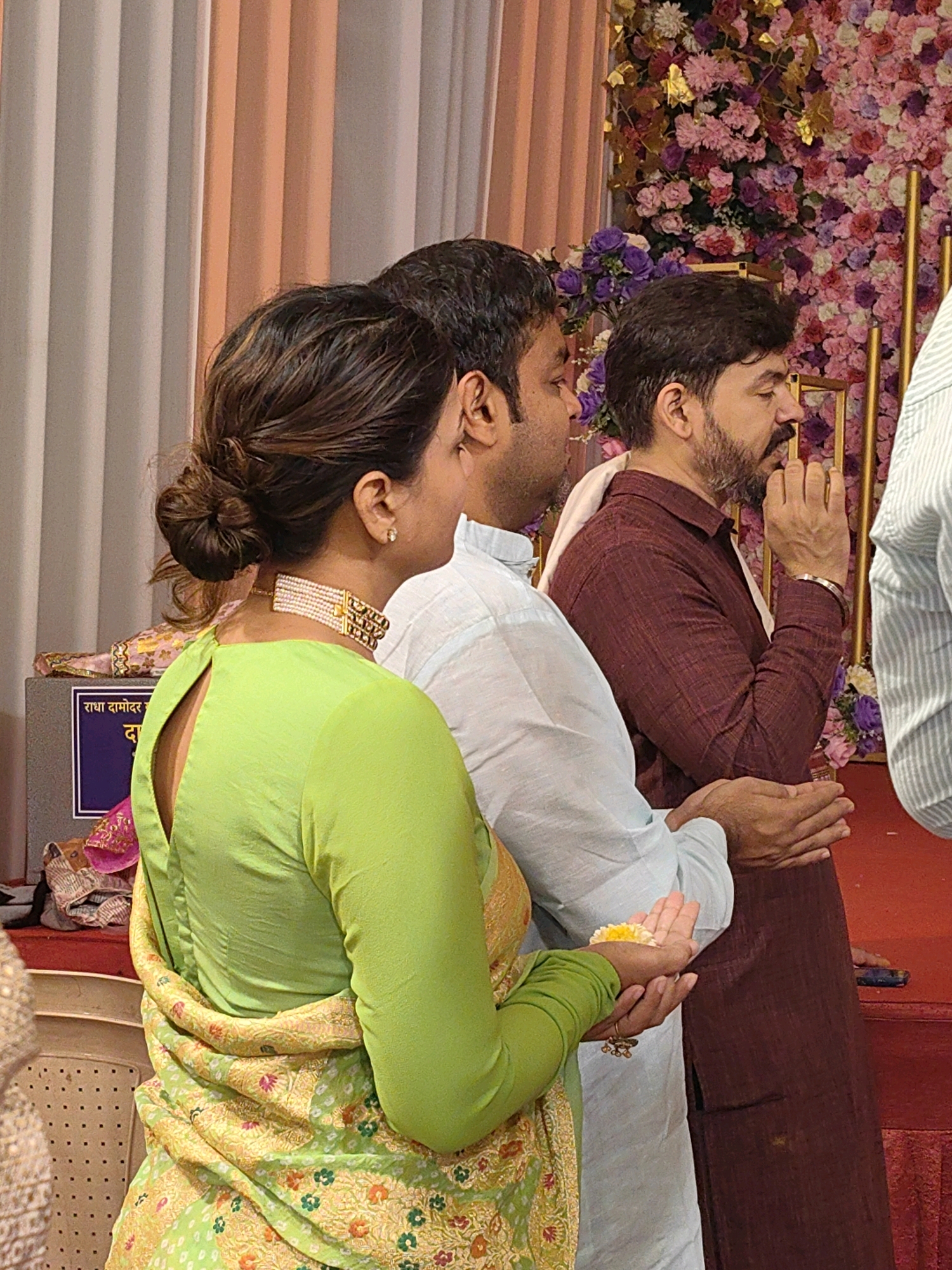A group of people are standing and facing right in front of a backdrop of pink and purple flowers. The person in the foreground is a woman wearing a bright green kurti and a yellow and green sari draped over her shoulders. She has her hair in a bun and is wearing a beaded choker necklace. Her hands are clasped together, holding a yellow flower. Next to her, a man in a light blue kurti is also facing right, with his hands clasped. Behind him, a man in a maroon shirt and pants is facing right, with his hands in a prayer-like gesture. The background features a decorative floral wall with vertical golden poles. A dark blue sign with white text is visible on a grey box to the left of the woman. The overall scene suggests a religious or cultural event taking place indoors, possibly in Mumbai, India, given the attire and floral decorations commonly seen in such settings.