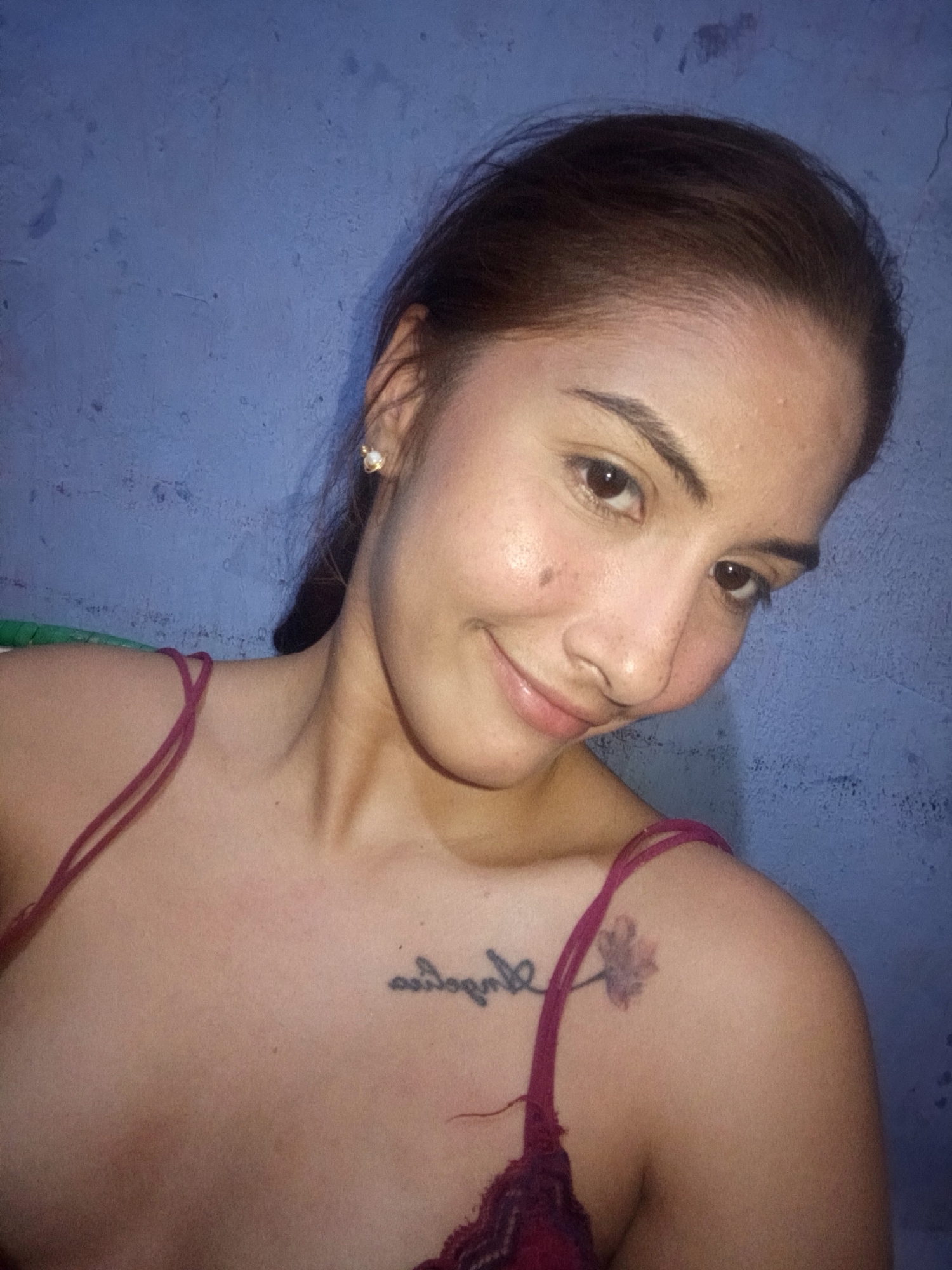 A fair-skinned woman with brown hair is posing indoors against a textured blue wall. She is wearing a camisole with maroon straps and lace trim. A tattoo of cursive writing and a flower is visible on her upper chest and shoulder. She is looking at the camera with a slight smile. An earring is visible in her left ear. The image provides no evidence to support its association with Juan Griego, Venezuela.