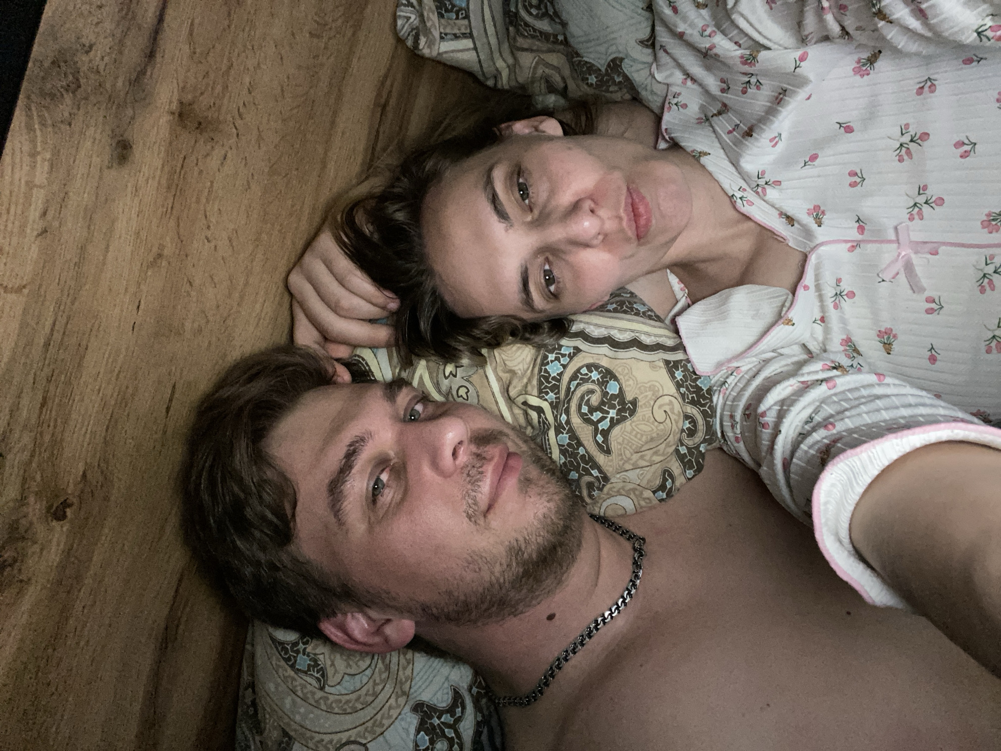 A couple, a man and a woman, are lying on a bed or mattress. The woman is on top wearing a white floral pajama top with pink trim. The man is shirtless and wears a silver chain. They are lying on patterned bedding. There is a wooden surface to the left, perhaps a floor or wall. It is impossible to determine the exact location in Chaikovsky, Russia from the image.