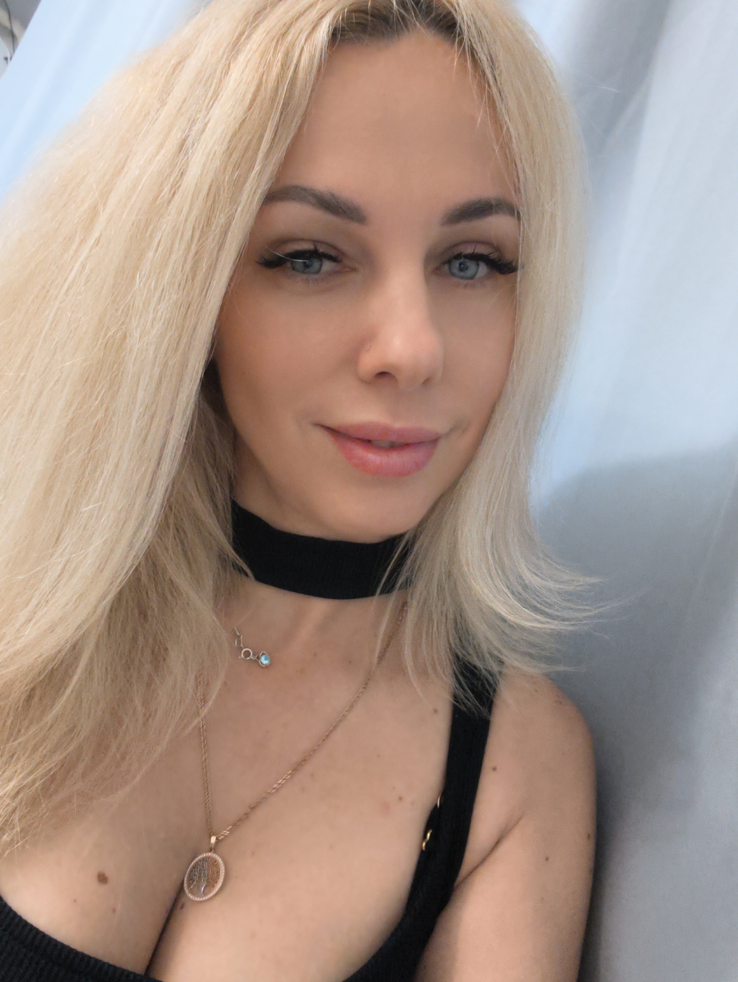 A close-up shot features a fair-skinned woman with platinum blonde hair and blue eyes. She is wearing a black choker and a longer gold necklace with a circular pendant depicting a tree. Her attire consists of a black top with thin straps. The background appears to be a light blue wall or curtain. There is no indication in this image that the photo was taken in Sevastopol, Ukraine.