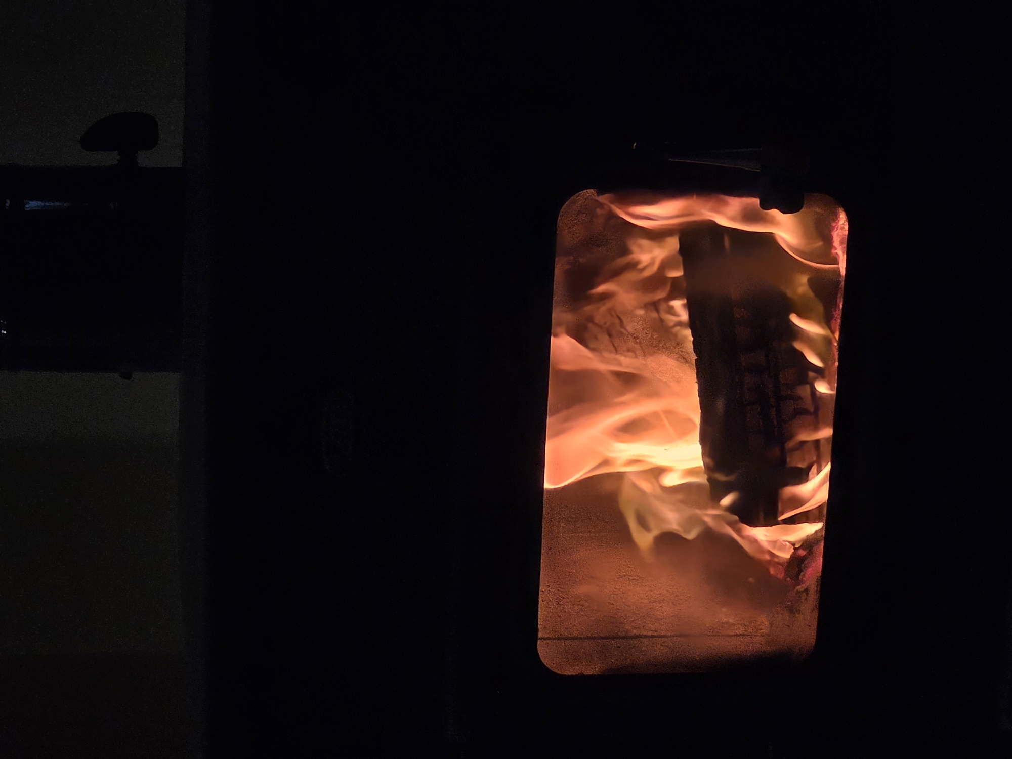 A low-light, close-up shot depicts a fireplace or wood stove with a rectangular window filled with flames. A dark piece of wood is burning inside the stove. The stove appears to be made of black metal. To the left of the stove is a shadowy, indistinct object or piece of furniture. No people are visible. There is no information to describe it is in Dulovo, Bulgaria.