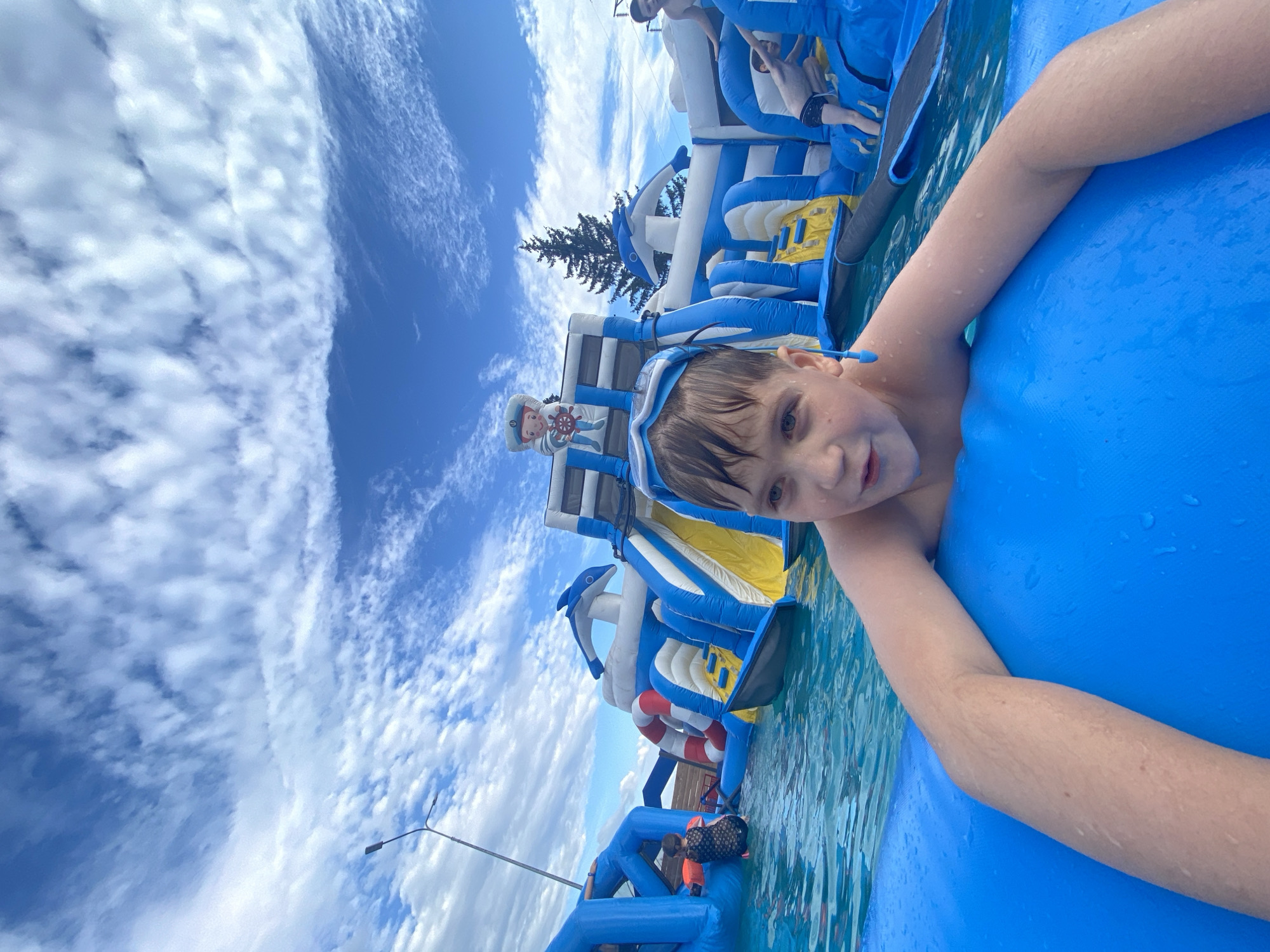 A young boy is shown in the foreground of this image, floating on a bright blue inflatable raft in a body of water. He is wearing goggles on his forehead and has wet hair. His right arm is extended out of the water, resting on the raft. The background features a large, inflatable water park with slides and climbing structures, predominantly in blue and white with yellow accents. There are other people visible on the water park, including one person dressed in a sailor costume at the base of a large slide. The sky above is a vivid blue, filled with scattered white clouds, suggesting it is a bright, sunny day. The overall scene depicts a fun recreational activity at an aquatic park. The location is Votkinsk, Russia.
