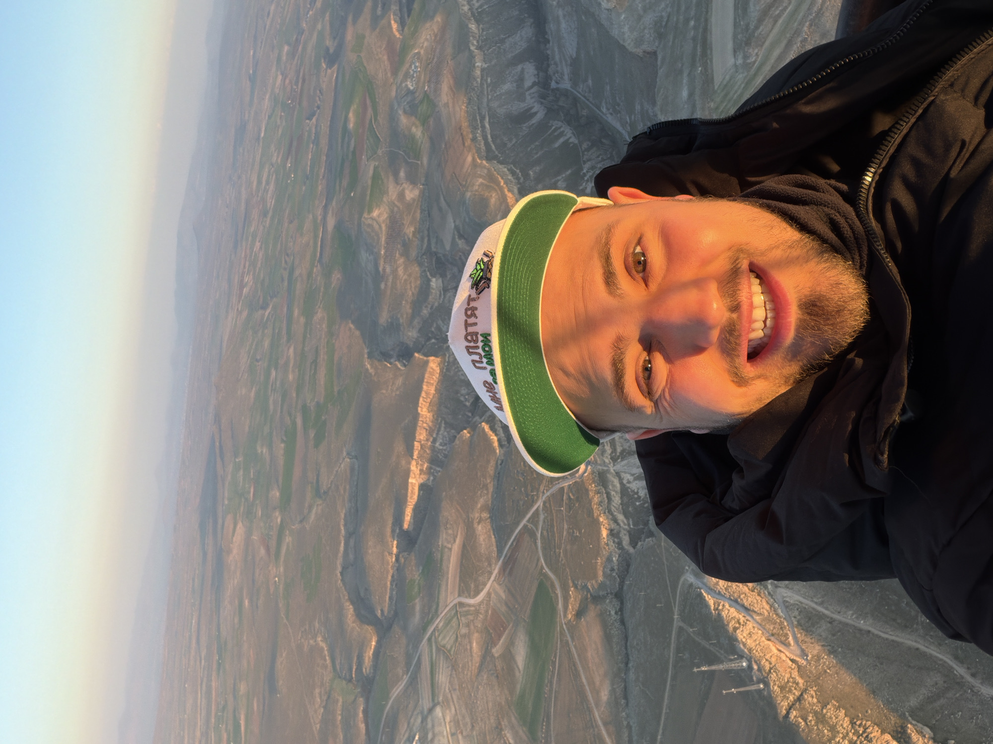 A selfie captures a smiling man against an expansive aerial view of a rugged, arid landscape, likely taken during sunrise or sunset. The man's face and upper torso fill the right side of the frame, illuminated by warm, golden light. He sports a beard and a wide, joyful smile, revealing his teeth. He is wearing a black hooded jacket and a white baseball cap with a green brim.
The text on his cap is in Cyrillic, reading "ПЛАТЯТ МОИ".
The background reveals a vast, undulating terrain, characterized by valleys, hills, and winding paths or roads. Scattered patches of green suggest agricultural fields amidst the predominantly earthy and brownish tones of the landscape. The golden hour light casts long shadows across the ground, emphasizing the contours of the land. In the distance, the horizon shows a light blue sky fading into a hazy, lighter hue. Some man-made structures, possibly utility poles, are visible in the lower right background. The high vantage point, combined with the distinctive geology, suggests a location like Yeşilhisar, Turkey, possibly observed from a hot air balloon. The clear weather allows for excellent visibility of the panoramic scene.