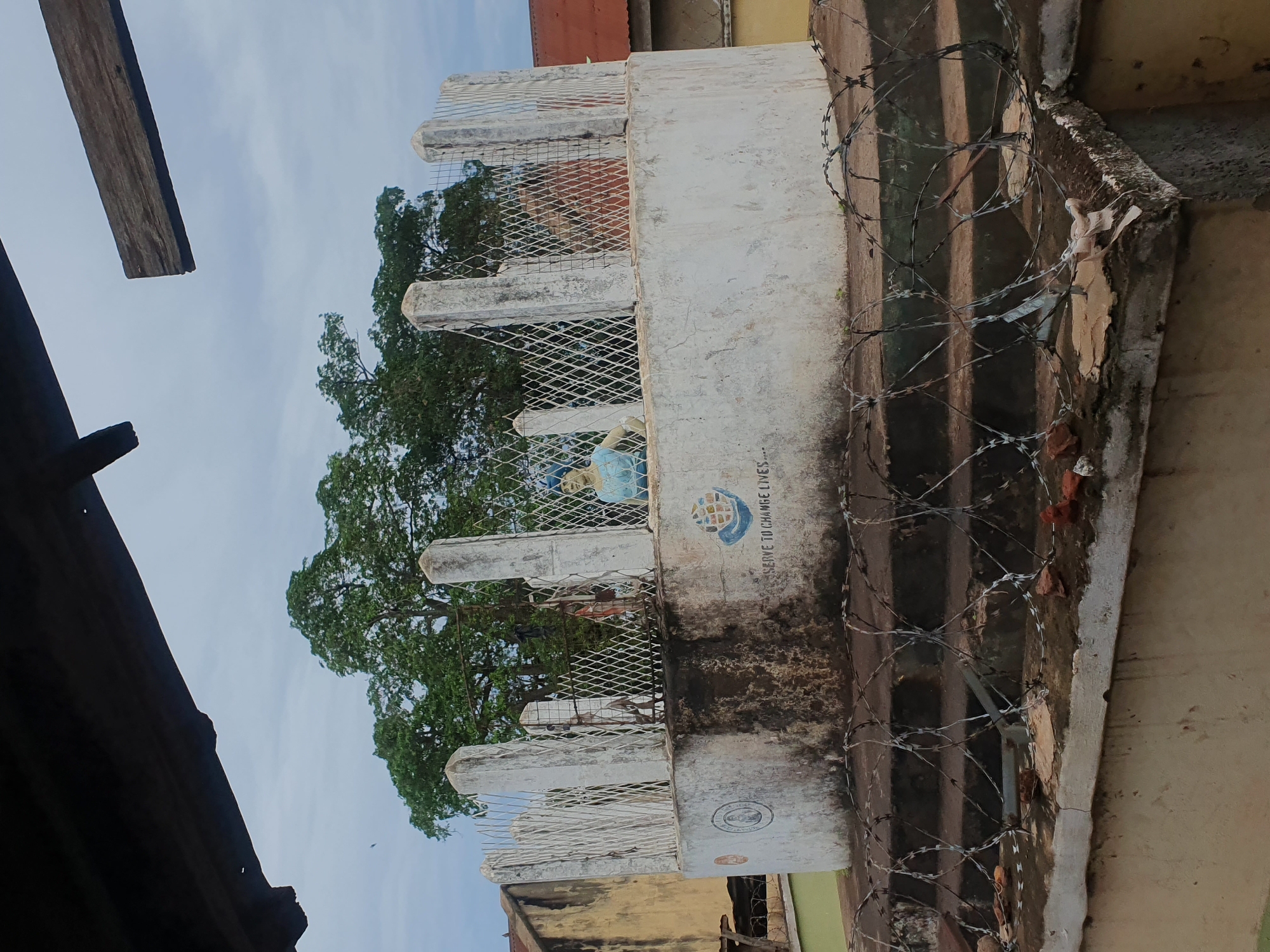 This image, captured in the city of Idah, Nigeria, depicts an outdoor scene under an overcast sky. The foreground is dominated by coils of razor wire (concertina wire) resting on a weathered concrete ledge, interspersed with small fragments of reddish-brown debris and light-colored refuse. Behind this wire, dark concrete steps are partially visible.
The central subject is a circular or semi-circular concrete structure, approximately shoulder height, exhibiting extensive dark staining and weathering. Mounted atop this structure is a fence composed of multiple vertical, rectangular concrete posts with a diamond-patterned metal mesh infill, showing signs of rust. Inside this enclosure, on the right, stands a light blue-colored statue of a human figure, partially obscured by the mesh and facing towards the left. The concrete wall bears a faded circular emblem on its left side and a prominent blue globe-like logo on its right, beneath which partially visible text reads "...SERVE TO CHANGE LIVES...".
The background features a dense canopy of large green trees, with parts of older buildings visible on both the left and right sides. The left background shows a portion of a light-colored building with a dark, possibly corrugated, roof. On the far right, a building with a reddish-brown corrugated roof is partially visible. Overhead, framing the top of the image, are dark, irregular shapes, likely parts of an architectural overhang or wooden beams, with a detached or broken wooden plank extending from the right. The sky is a uniform light gray, indicating cloudy conditions.