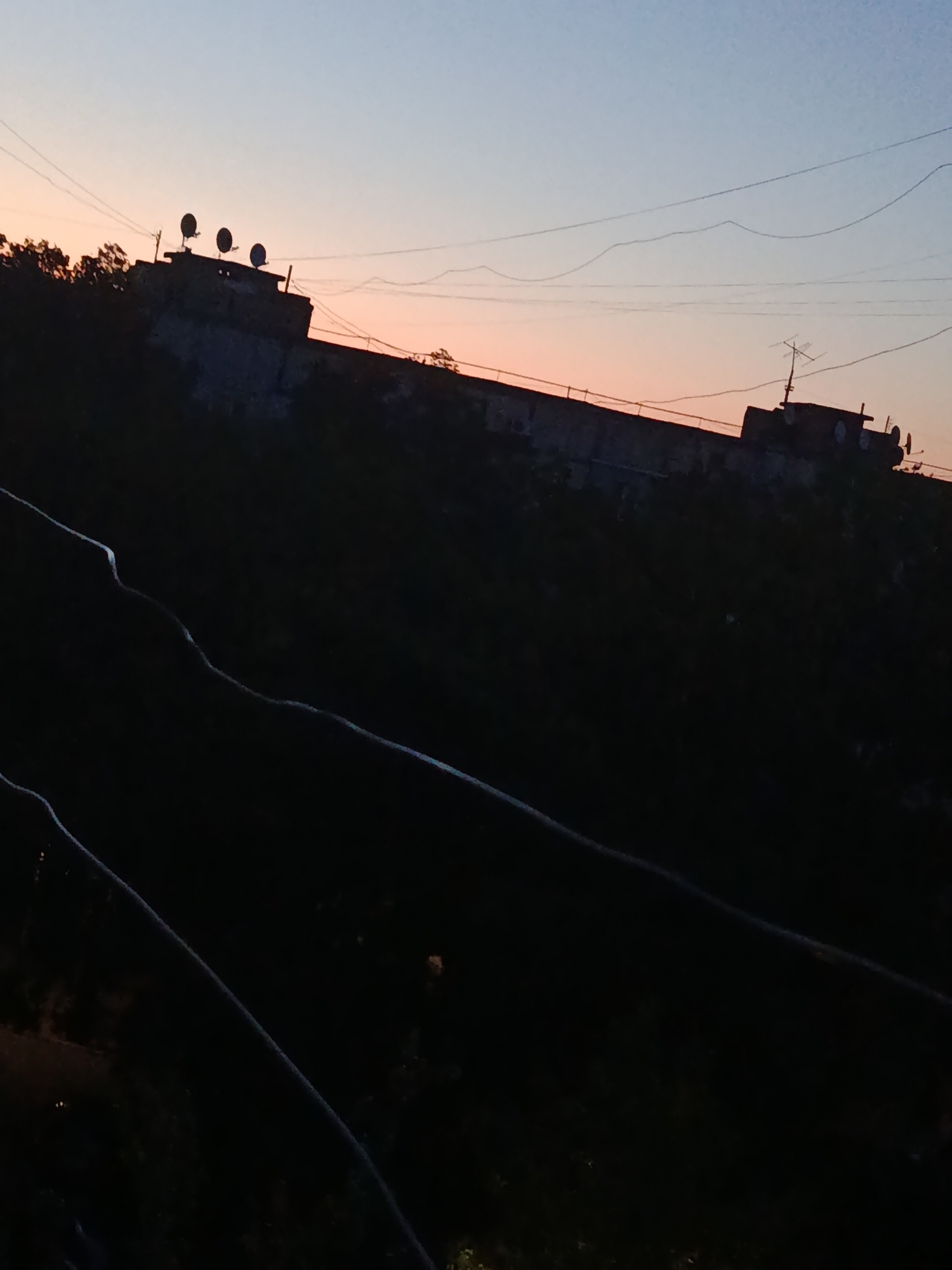 The image depicts a low-angle shot of a long, dark, silhouetted structure against a twilight sky. The structure appears to be a building or a part of a larger complex, possibly industrial, with several dish-like antennas on its roof. The sky is a gradient of soft blues and oranges, indicating either sunrise or sunset. There are thin, dark lines crisscrossing the sky, likely power lines or telephone wires. The foreground is almost entirely black, obscuring any details of the immediate ground or surroundings. The tilted perspective and the darkness suggest a casual, possibly handheld, photograph taken at dusk or dawn. The specific location in Cherkaske, Ukraine, is not visually identifiable from the provided image itself.