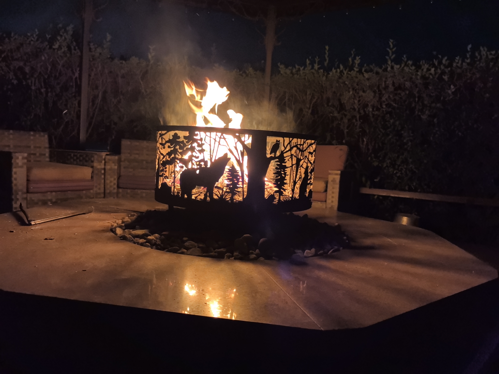 The image depicts a nighttime scene centered around a fire pit in what appears to be a backyard or patio area. The fire pit itself is a rectangular metal enclosure with a decorative silhouette design featuring a wolf, trees, and an owl. Flames are visible inside the fire pit, casting a warm glow on the surrounding area. The fire pit rests on a circular stone border, which sits on a large, flat, dark-colored surface, likely a patio table or platform. The surface reflects the light from the fire, creating bright, shimmering patterns.
In the background, there are glimpses of outdoor furniture, including brick structures with cushioned seats. Beyond the immediate seating area, dense foliage and trees form a dark backdrop against the night sky, which has a few faint white specks that could be stars. The overall atmosphere is one of a cozy outdoor gathering or relaxation space at night.
The city of Taldykorgan, Kazakhstan, is not depicted or suggested in the provided image. There are no visible landmarks, signs, or environmental features that would indicate this specific location. The scene is generic to many outdoor settings with a fire pit.
