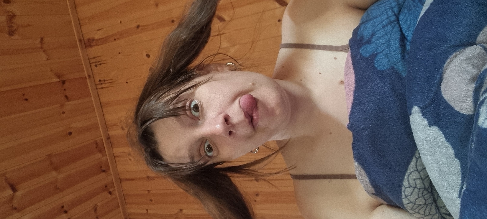 The image shows a woman with brown hair in pigtails sticking her tongue out. She is wearing a brown tank top and is partially covered by a blue and grey patterned blanket. The background consists of wooden planks. The location of the city of Dunnae-myeon, South Korea is not identifiable.