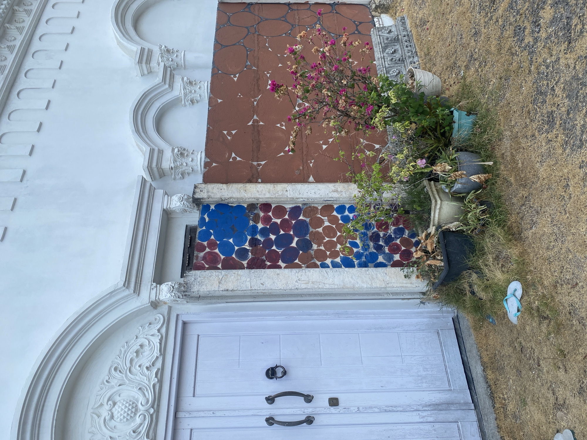 In Banjarbaru, Indonesia, a pale blue, weathered double door stands to the left, featuring ornate black door handles and a smaller black doorknob. To its right, a column of rough stone separates it from a section of wall adorned with colorful circles in blue, red, and brown. Further to the right, a rust-colored wall displays a repeating pattern of white triangular shapes within circular outlines. A bougainvillea plant with pink and faded blossoms grows in front of this wall, alongside several potted plants on dry, yellowed grass. A single light blue flip-flop lies on the ground near the door. The building's facade exhibits decorative white architectural details, including arched structures and carved ornaments.