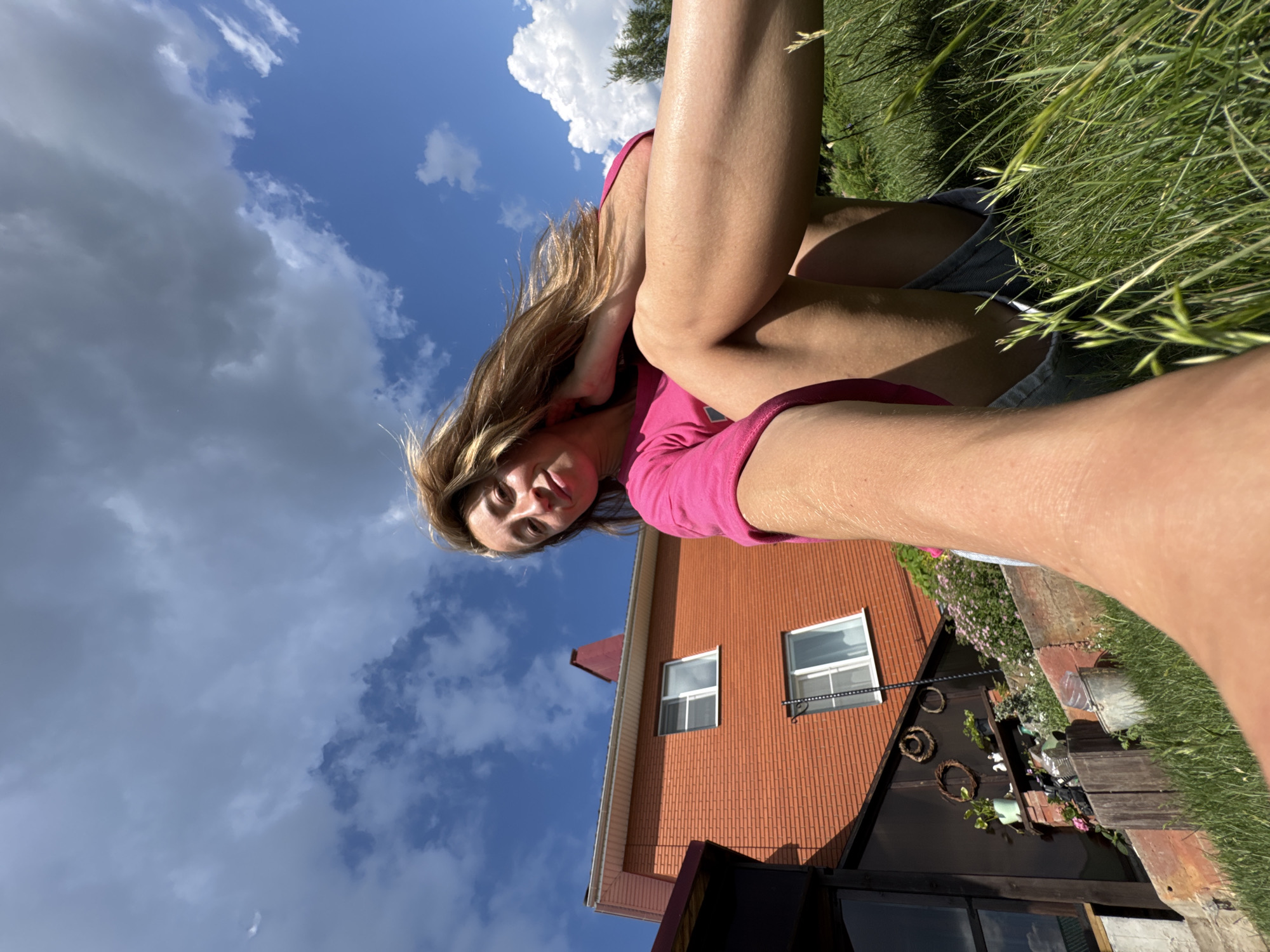 The image depicts a young woman sitting on a grassy hill. She is wearing a bright pink t-shirt and denim shorts. Her hair is blonde and flowing in the wind. In the background, there is a two-story brick house with a red roof and several windows. The sky is blue with scattered clouds. The scene appears to be set in a rural or suburban area, possibly in the city of Прикамский, Russia, given the architectural style of the house and the lush green landscape. The woman is looking towards the camera and smiling. The photo is taken from a low angle, emphasizing the sky and the woman's presence in the natural environment.
