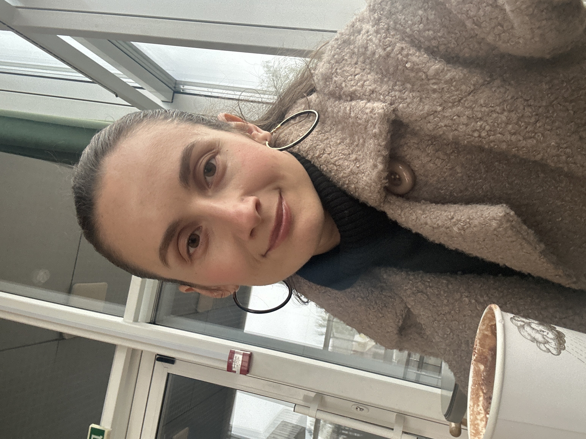 This selfie, taken in Kryvyi Rih, Ukraine, features a woman smiling gently at the camera. She has dark hair pulled back, showcasing large, dark hoop earrings. She is dressed in a dark, high-necked top underneath a light brown or beige textured boucle coat, with a visible button on the lapel. Her expression conveys a relaxed and pleasant mood.
The setting appears to be indoors, characterized by large, modern windows that provide natural, diffuse lighting, suggesting an overcast or cloudy day outside. Architectural elements like window frames and ceiling structures are visible in the background. In the lower right corner, a white paper coffee cup with an indistinct decorative pattern, possibly floral or leaf-like, is partially visible, indicating she might be enjoying a drink.
Notable details in the background include a red fire alarm box mounted on a window frame, with the word "FIRE" faintly discernible. A partially obscured green sign, possibly indicating "EXIT" or a similar directive, is also visible near a door in the lower left section of the image. The overall atmosphere is tranquil, suggesting a moment of quiet leisure.