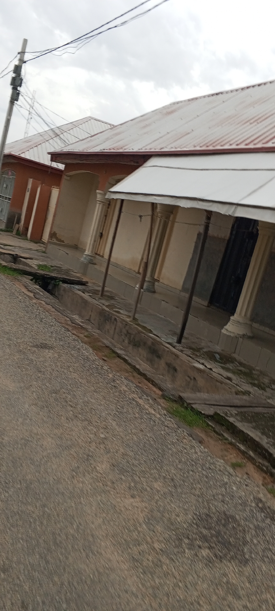 This is a photograph of a residential street in Jalingo, Nigeria. The street is paved with asphalt and has a drainage ditch running along its side. On the right side of the street, there is a row of buildings with corrugated metal roofs. The buildings appear to be old and in need of repair. The sky is cloudy and overcast. There are no people or animals visible in the photograph.