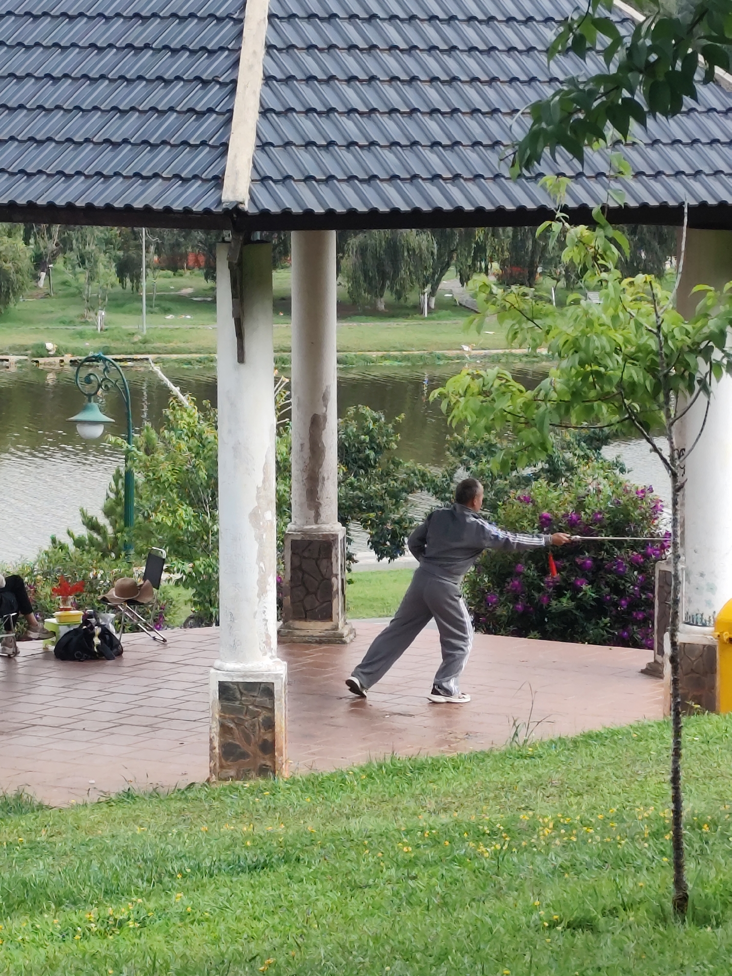 In Da Lat, Vietnam, a person in a gray tracksuit performs a martial art with a long staff inside a gazebo on a stone-tiled platform. The gazebo has a dark tiled roof supported by white columns with decorative stone bases. The person is facing right, extending the staff towards a bush with purple flowers. A small tree stands near one of the columns. In the background, a lake is visible with trees lining its banks. A lamppost and some chairs are seen on the left side of the gazebo. The foreground is a patch of green grass with small yellow flowers.