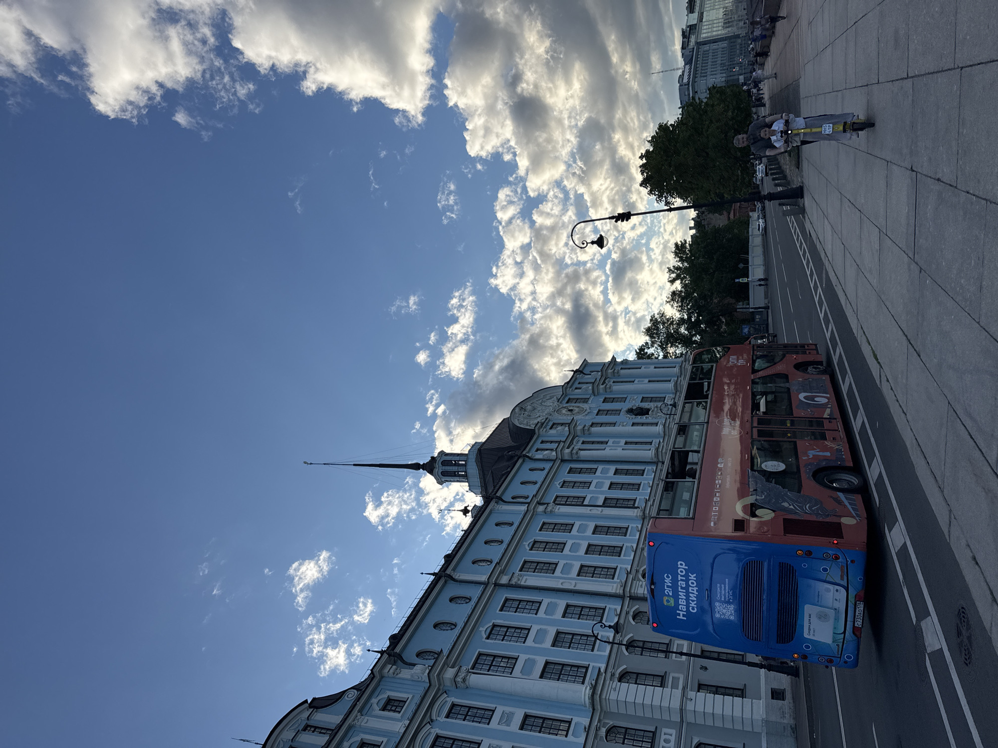 In Saint Petersburg, Russia, a double-decker tour bus is captured from the rear, driving on a street next to a light blue, ornate building. The bus, predominantly blue and red, displays Russian text and a QR code on its rear. A father and child are seen on the right side of the image, riding an electric scooter on a paved pedestrian walkway. The father is steering, with the child positioned in front of him. The backdrop features a clear blue sky with scattered clouds, trees, and other city buildings. The overall scene depicts a city street with urban transportation and pedestrian activity under a partly cloudy sky.