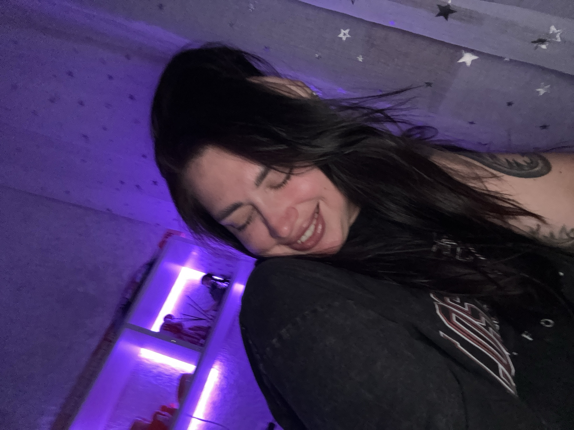 The image shows a woman with dark hair, smiling with her eyes closed. She is wearing a black t-shirt and is leaning against a curtain with a star pattern. Behind her, shelves are lit with purple light. The scene appears to be indoors, possibly a bedroom, and the overall atmosphere is dimly lit with purple accents. There is no indication of the location within Tolyatti, Russia.