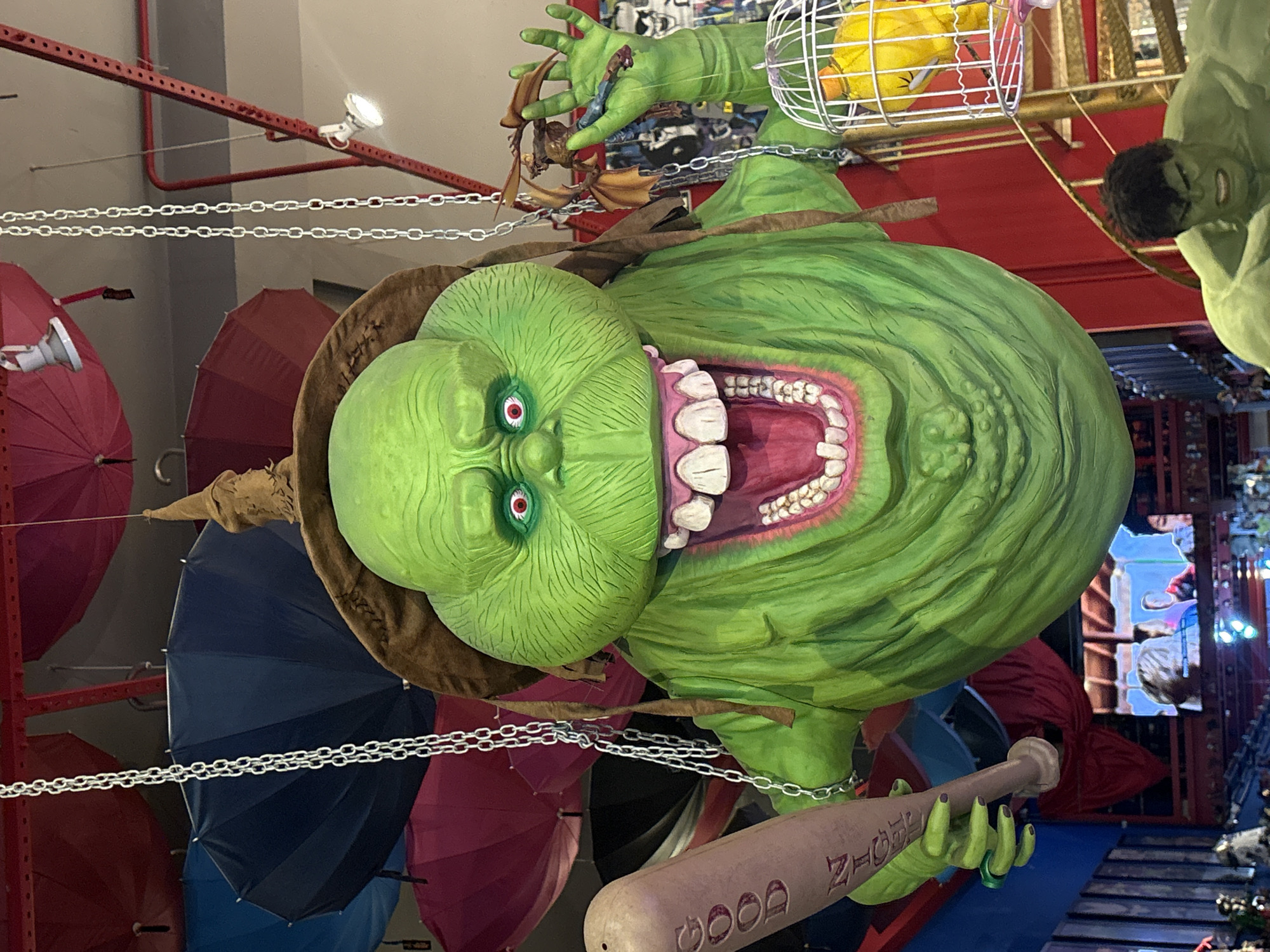 The image depicts a large, green, inflatable character resembling the Stay Puft Marshmallow Man from Ghostbusters, suspended from the ceiling by chains. The character wears a pointed hat and has a wide, toothy grin, red eyes, and visible wrinkles. It is holding a pink baseball bat in its left hand, with the word "GOOD" written in red on the bat. In its right hand, it holds a small, brown, winged creature. Behind the character, several colorful umbrellas are visible, along with a white birdcage containing a yellow bird. In the foreground, a figure of the Hulk is partially visible, with a green, muscular body. The background shows shelves stocked with various items, including what appear to be collectible figures and posters. The scene is indoors, likely a shop or exhibition, with a display of merchandise and promotional items. This scene is located in a store in the Bom Retiro neighborhood of São Paulo, Brazil.