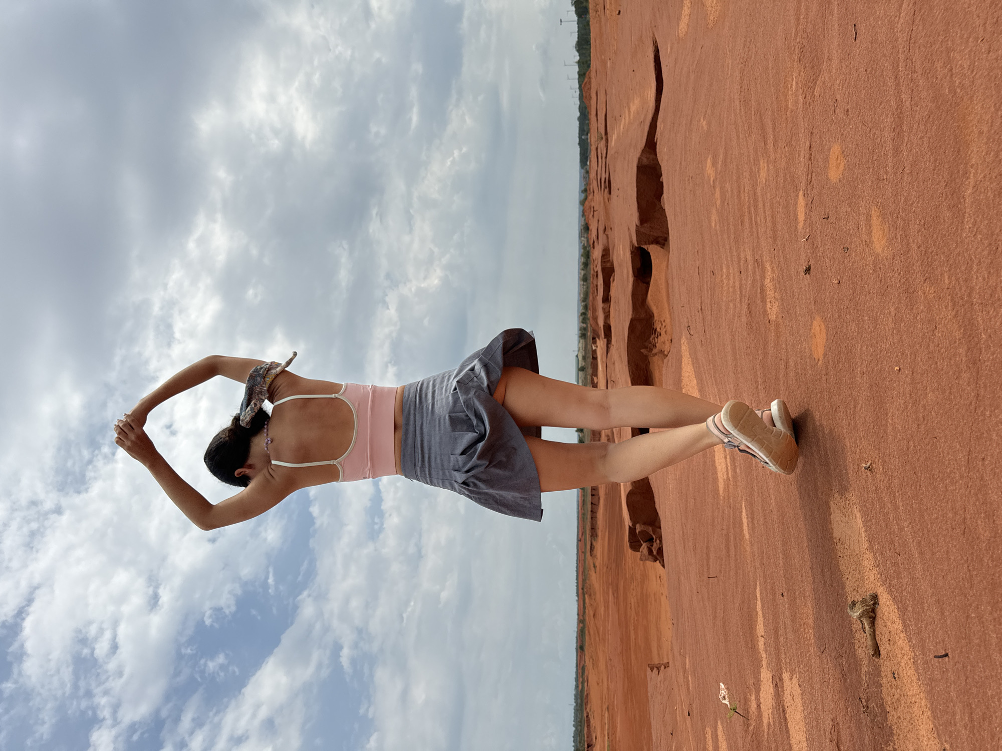 A person is viewed from behind, standing on red sand with a barren landscape stretching to the horizon under a cloudy sky. The person is wearing a pink crop top, a gray pleated skirt, and athletic shoes. Their arms are raised above their head, and their body is slightly turned to the left. The red sand landscape appears to be a desert-like environment with eroded formations. The overall scene suggests a remote, dry location. This location is the Red Sand Dunes in Mui Ne, Vietnam.